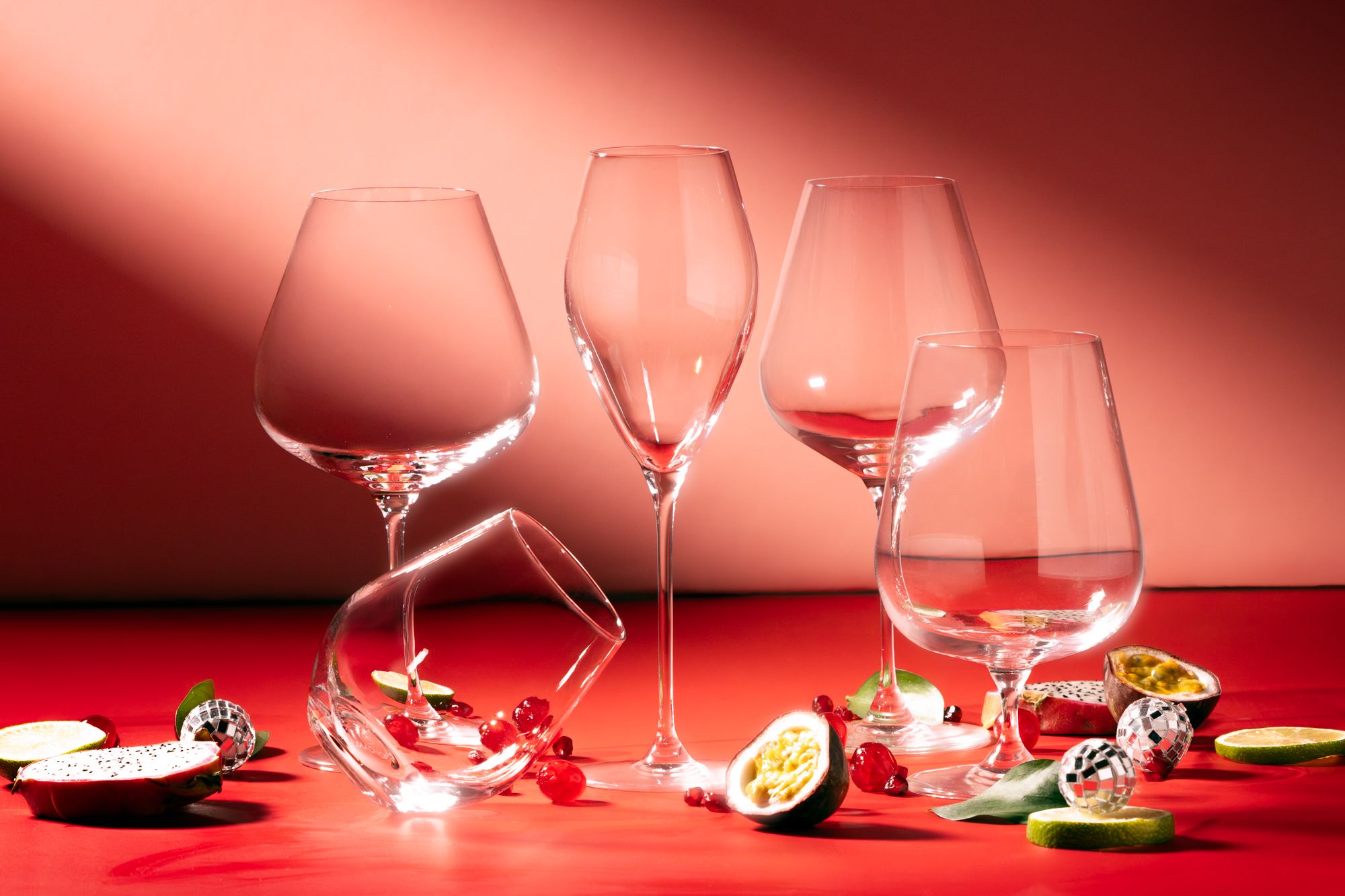 Elegante vinglass står på et rødt bord omgitt av eksotisk frukt og dekorasjoner mot en varm, rosa bakgrunn.