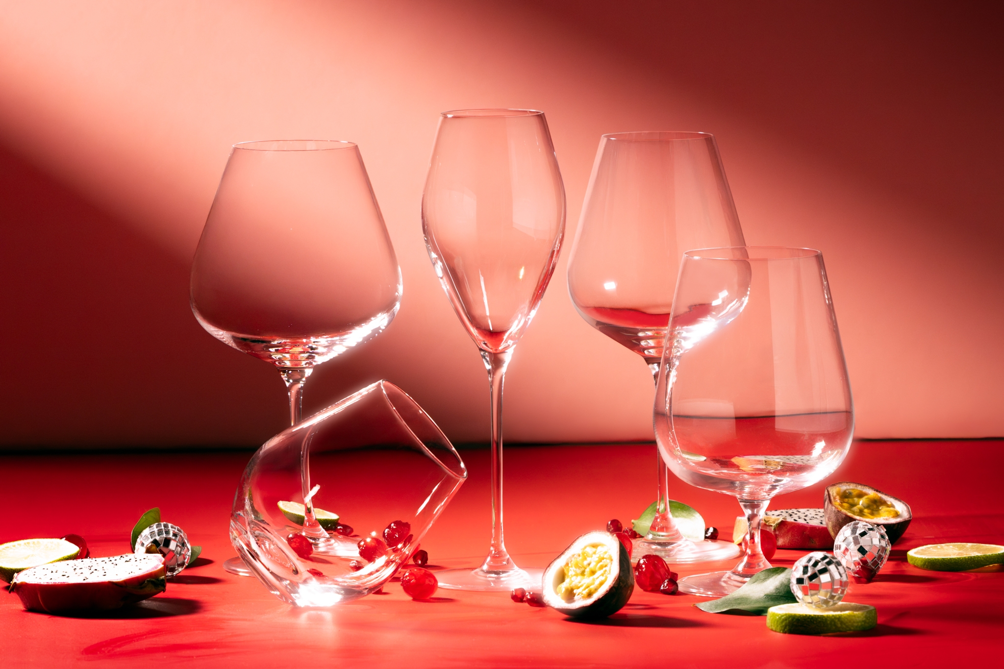 Elegante vinglass står på et rødt bord dekorert med pasjonsfrukt, lime og bær mot en varm, rosa bakgrunn.