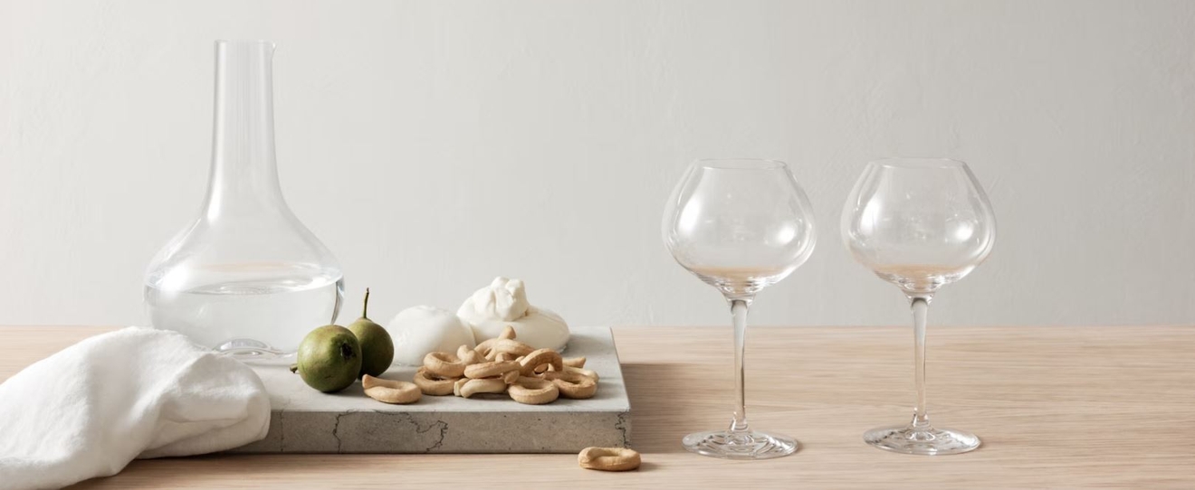 Minimalistisk borddekking med karaffel, to vinglass, småkaker og grønne frukter på lys treflate.
