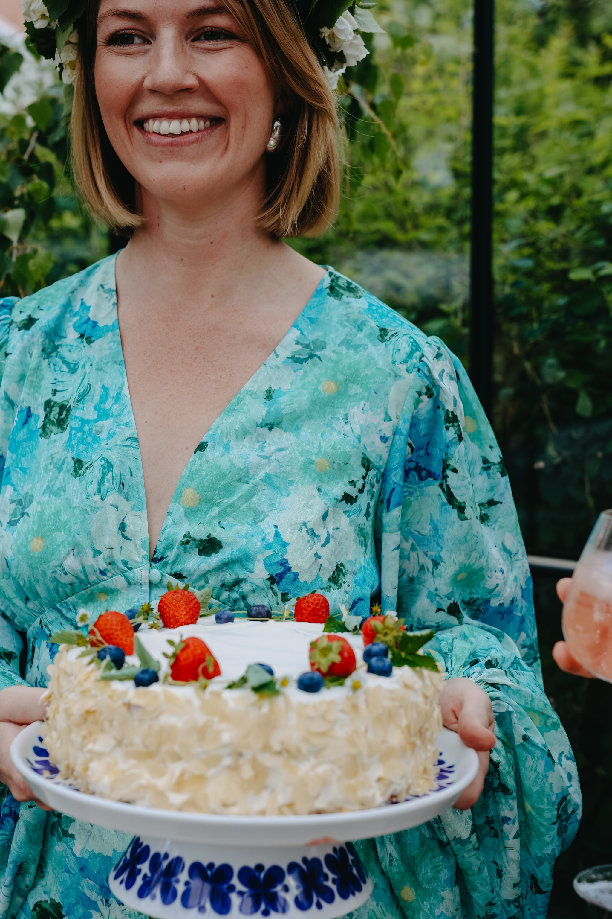 Leende kvinna i blommig sommarklänning håller en gräddtårta dekorerad med jordgubbar och blåbär utomhus.