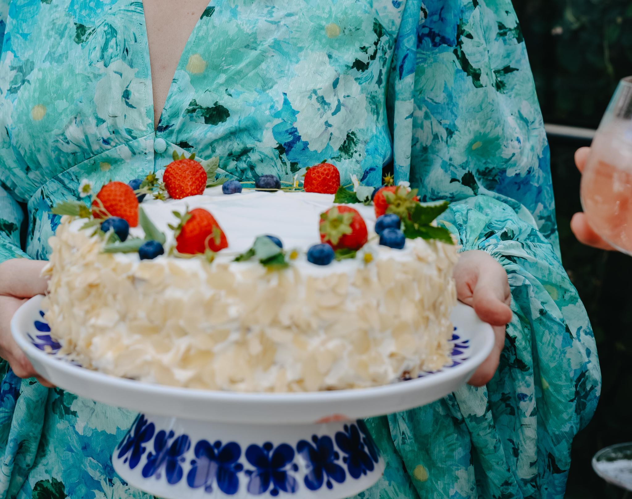 Leende kvinna i sommarklänning håller en gräddtårta dekorerad med jordgubbar och blåbär utomhus i grönskande trädgård.