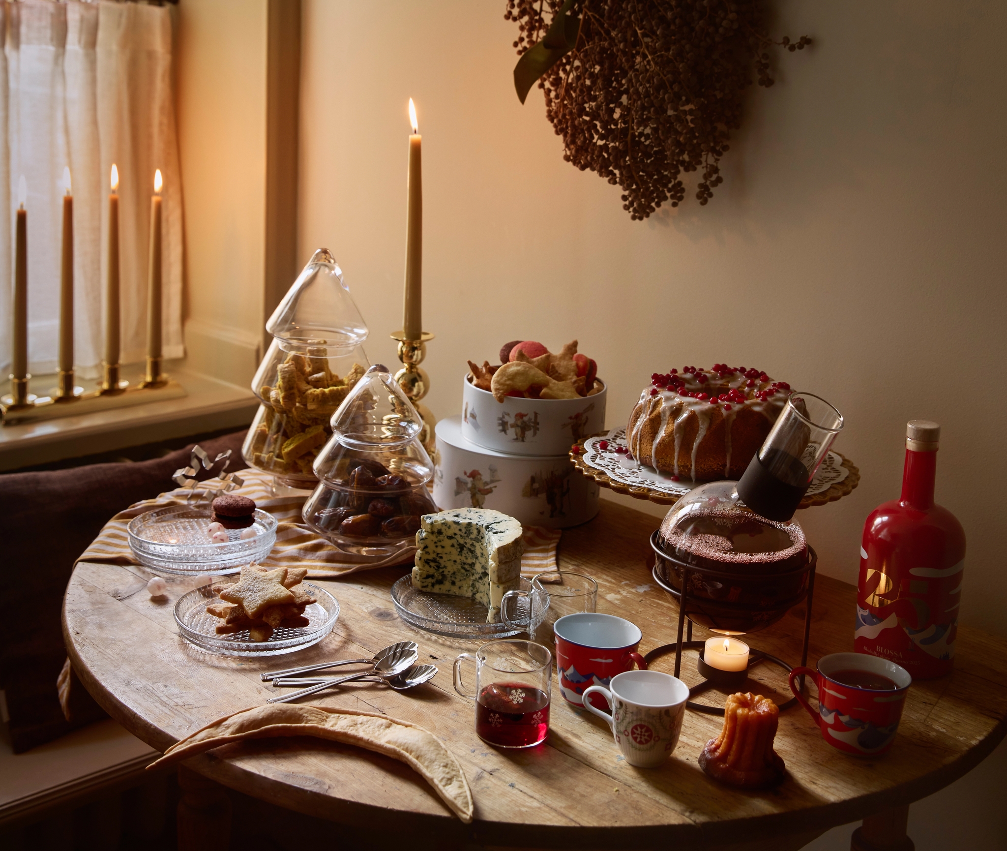 A rustic table filled with holiday treats like cakes, cookies in tree-shaped jars, cheese, and warm drinks, lit by candlelight in a cozy room.