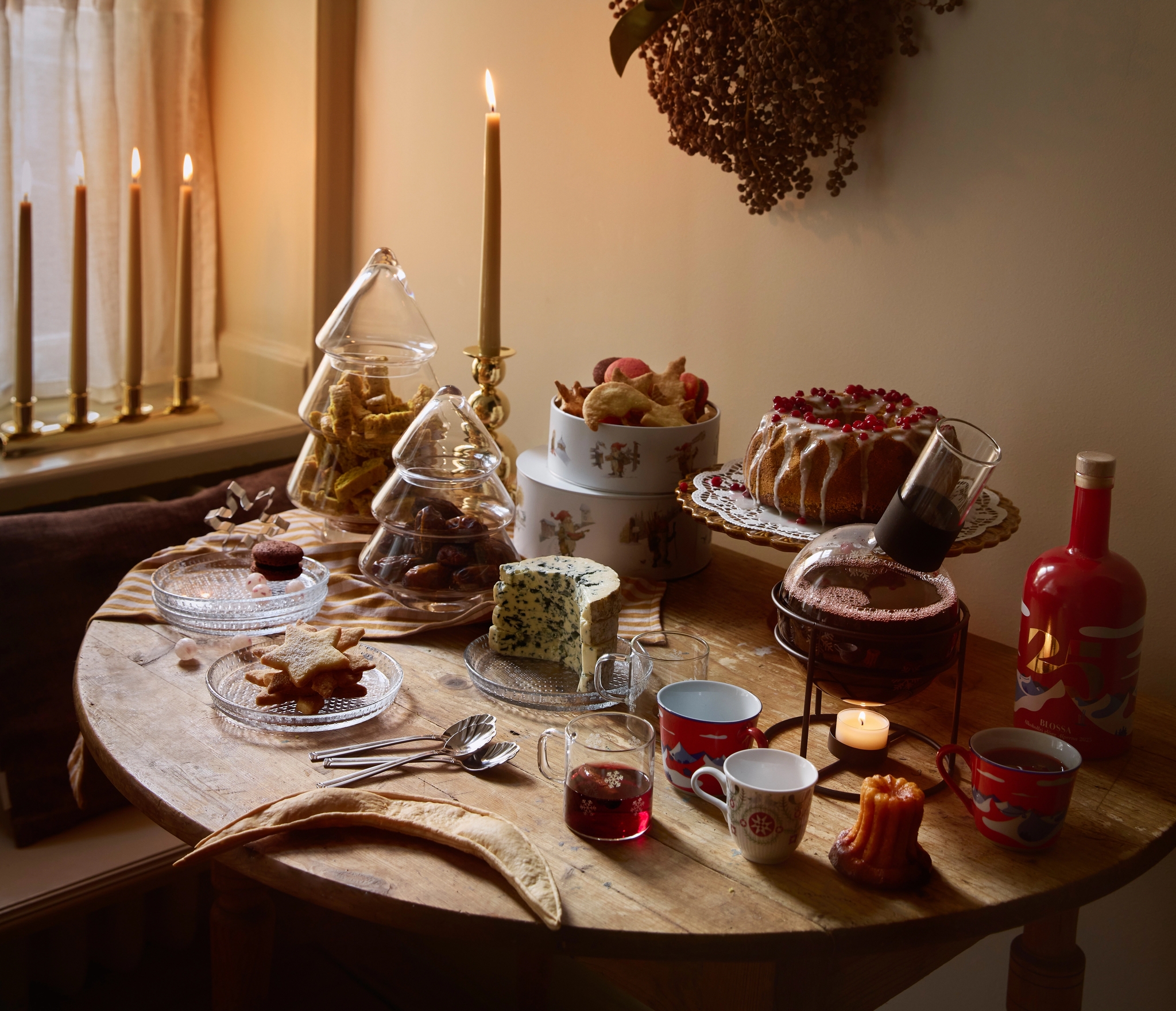 A cozy, candlelit festive table by a window, featuring cakes, cookies in tree-shaped jars, blue cheese, and drinks, with a rustic dried wreath on the wall.