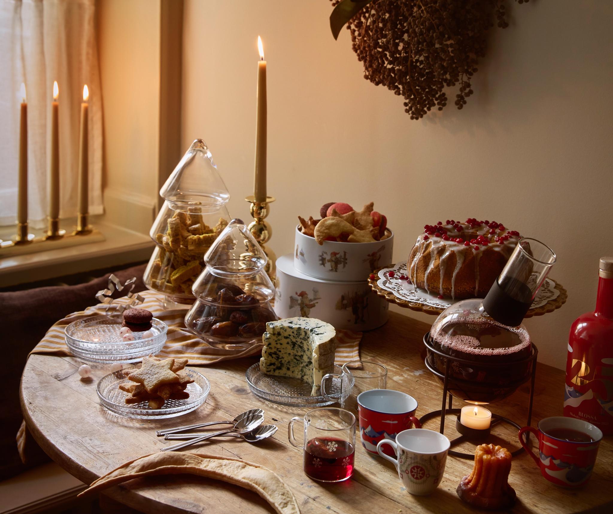 Juhlava, kynttilänvalossa loistava pöytä, joka on katettu naposteltavilla, jälkiruoilla ja juomilla ikkunan vieressä.