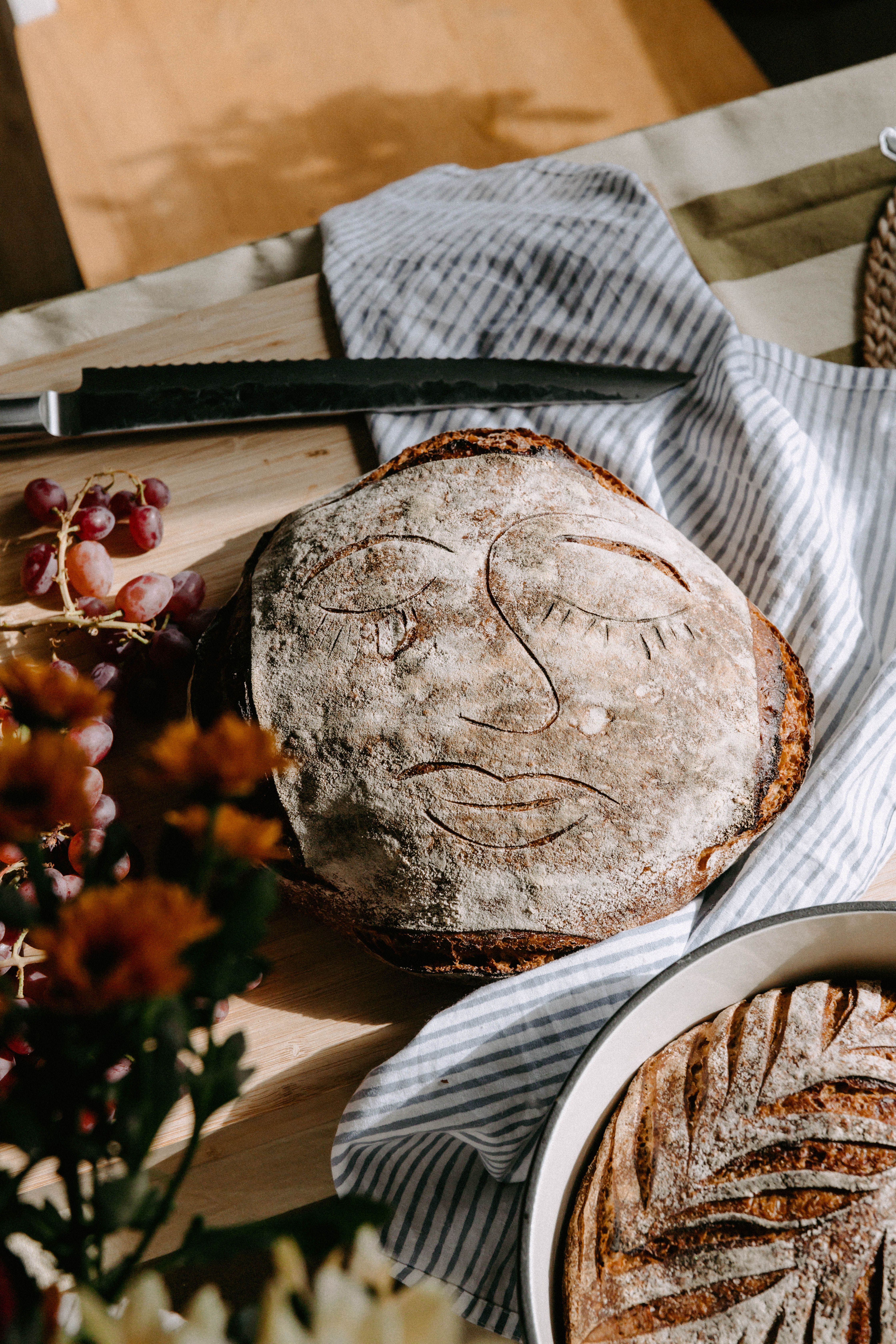 Rustikt surdegsbröd med ett konstnärligt ansikte inristat i skorpan ligger på ett köksbord med druvor och blommor runtomkring.