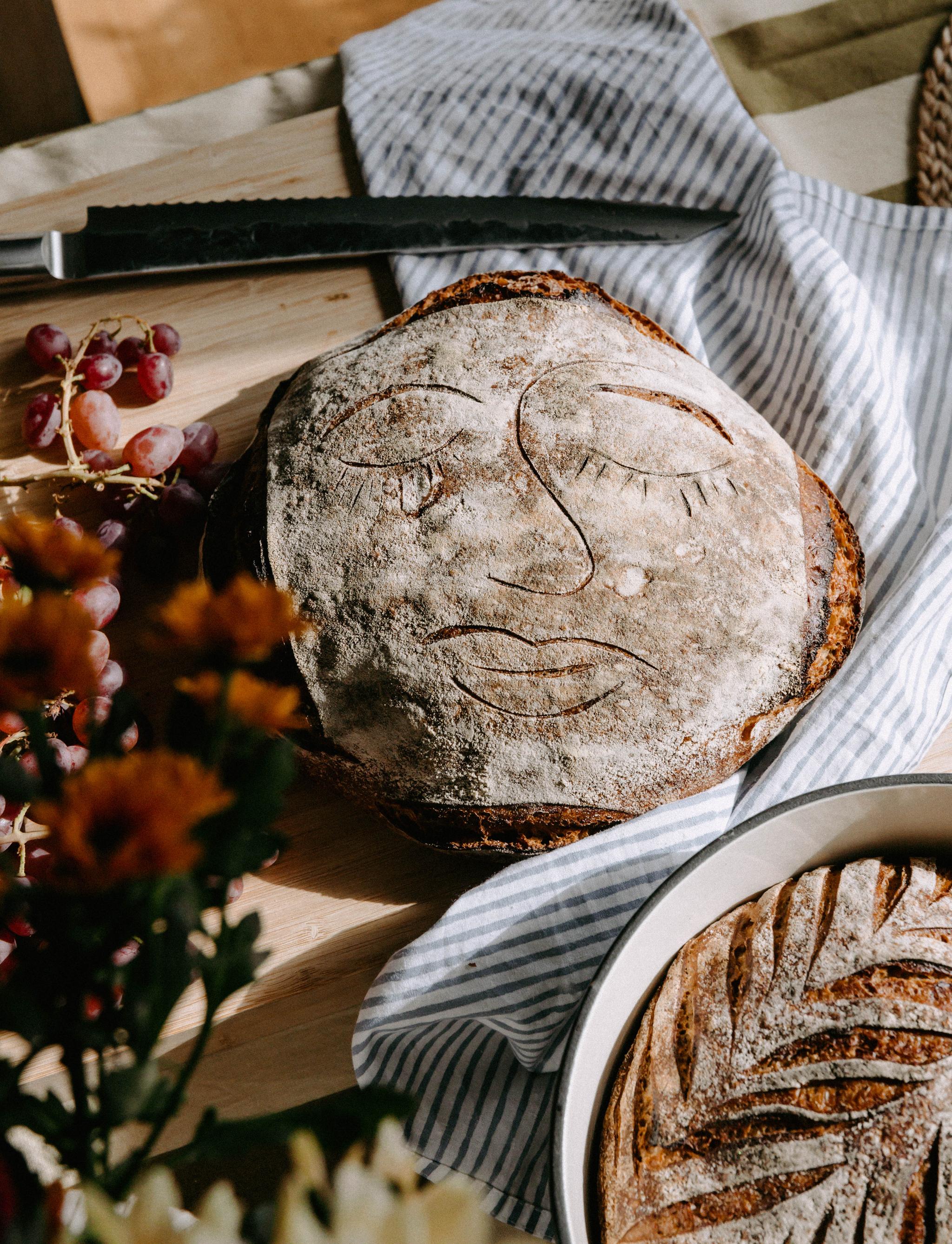 Rustikt surdegsbröd med ett konstnärligt ansikte inristat i skorpan ligger på ett köksbord med druvor och blommor runtomkring.