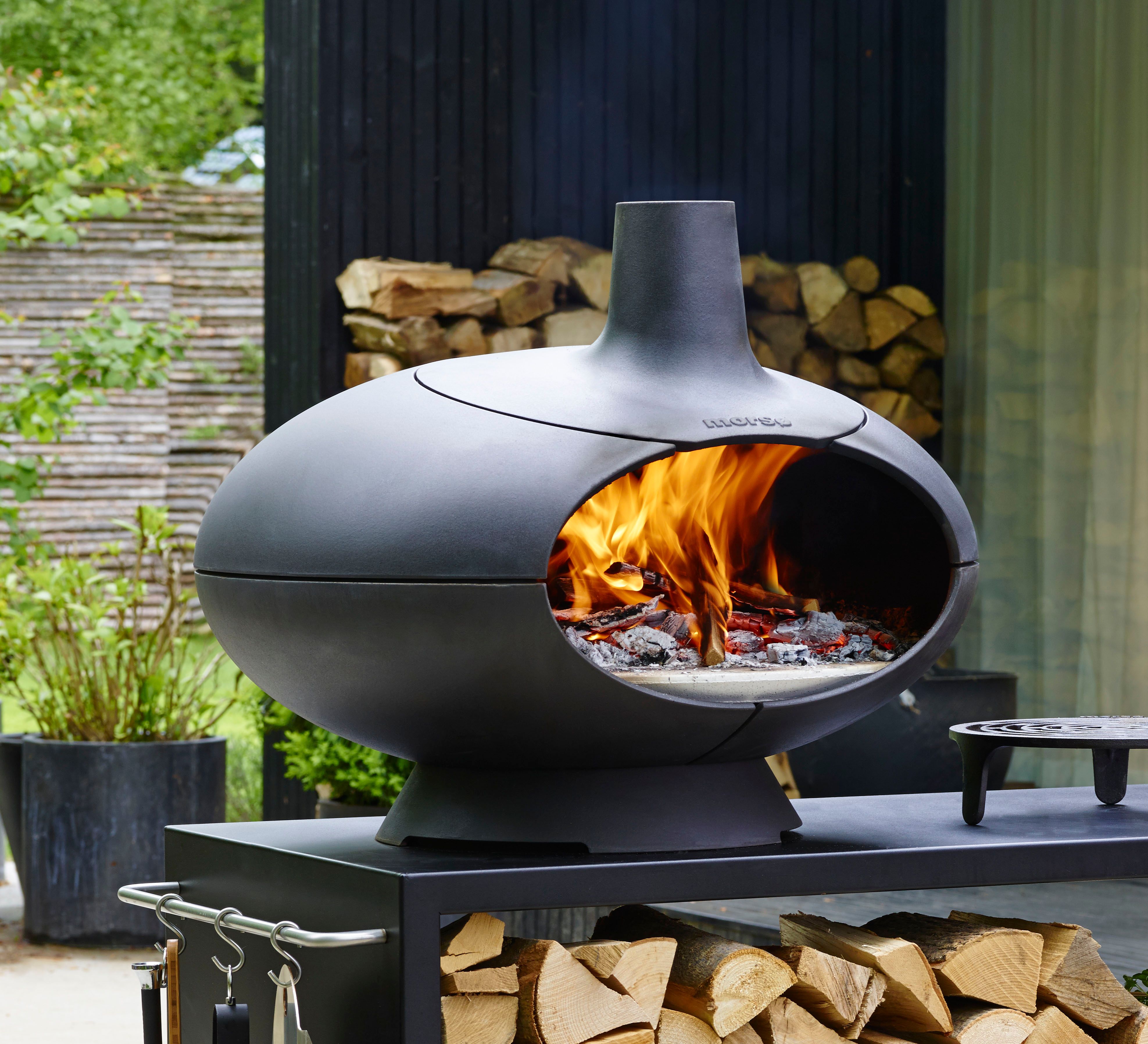 Black, egg-shaped outdoor pizza oven with a bright fire burning inside, on a stand with stacked firewood.