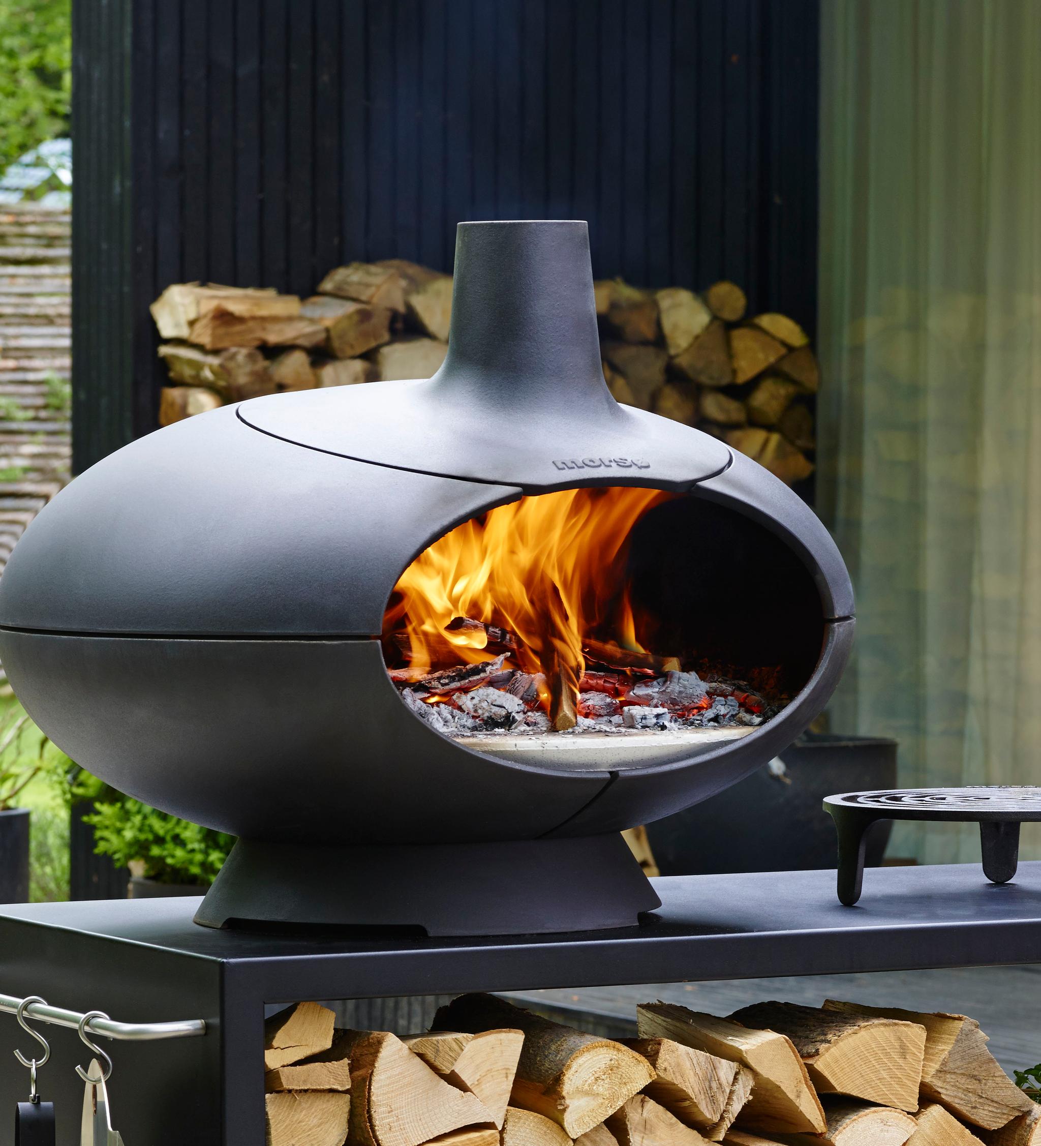 Black, egg-shaped outdoor pizza oven with a bright fire burning inside, on a stand with stacked firewood.