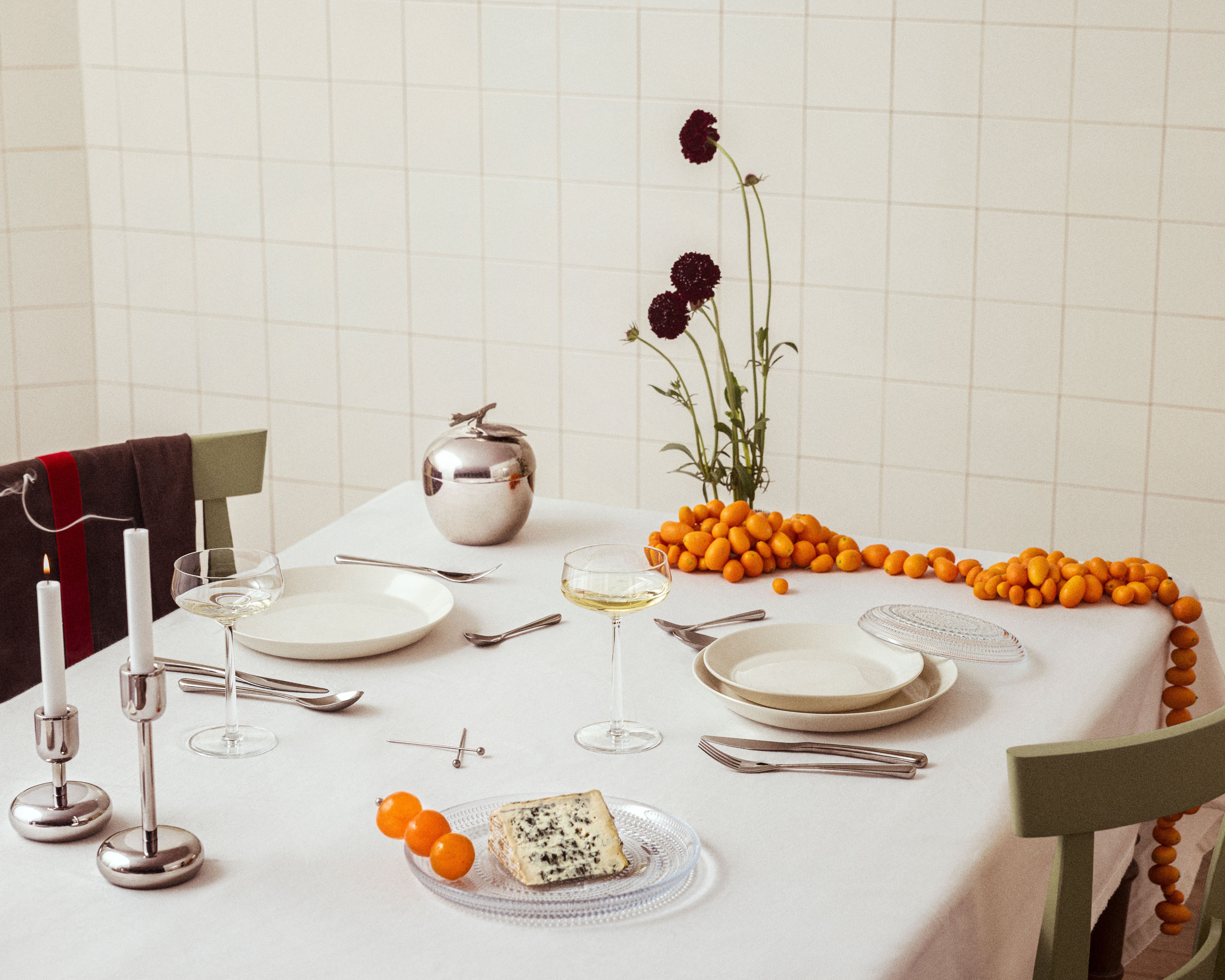 Ruokapöytä, jossa valkoiset lautaset, samppanjalasit, sytytettyjä kynttilöitä, hopeinen omenanmuotoinen rasia, tummanpunaisia ​​kukkia, sinihomejuustoa ja kumkvatteja valkoisella pöytäliinalla.