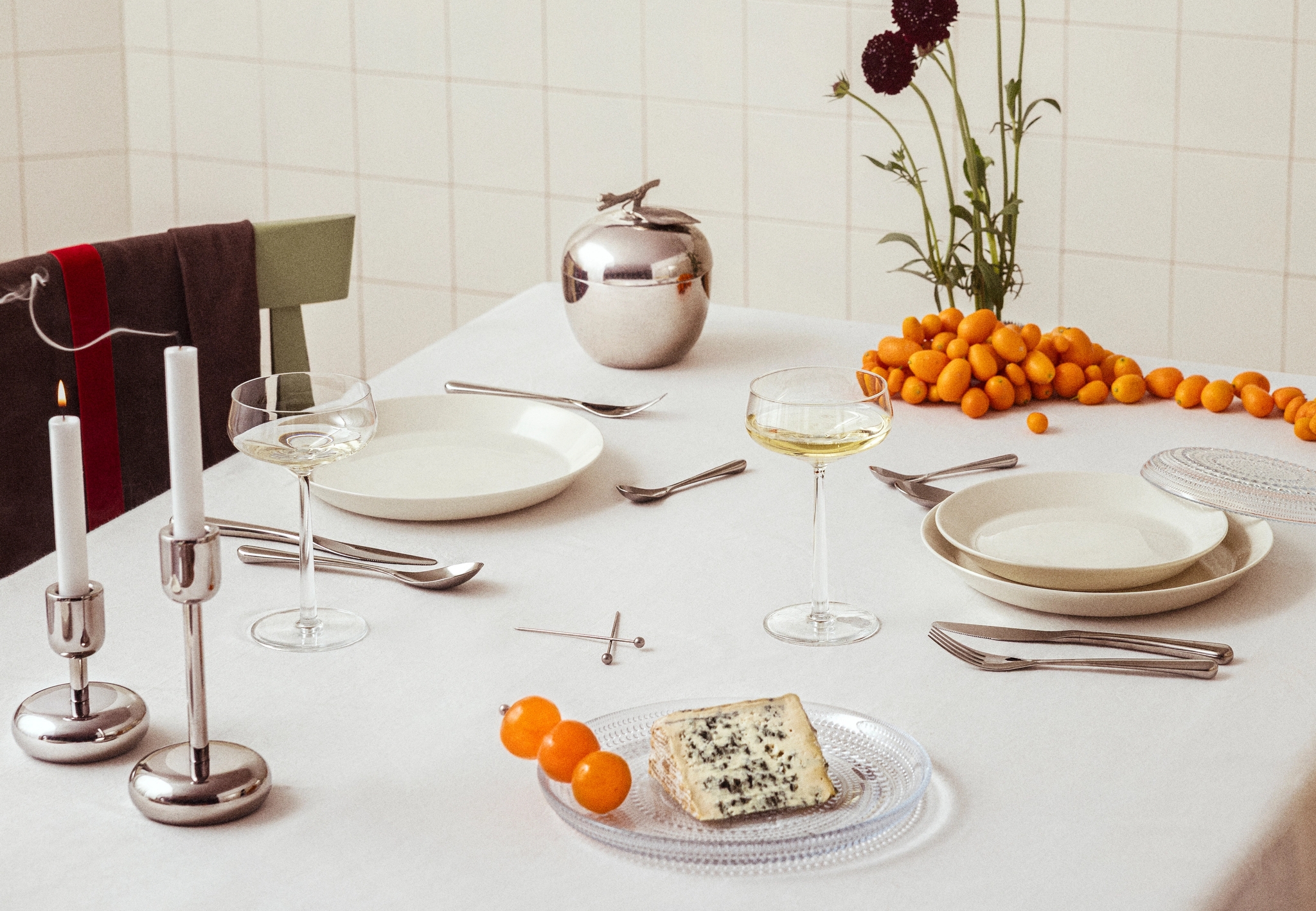 Ruokapöytä, jossa valkoiset lautaset, samppanjalasit, sytytettyjä kynttilöitä, hopeinen omenanmuotoinen rasia, tummanpunaisia kukkia, sinihomejuustoa ja kumkvatteja valkoisella pöytäliinalla.