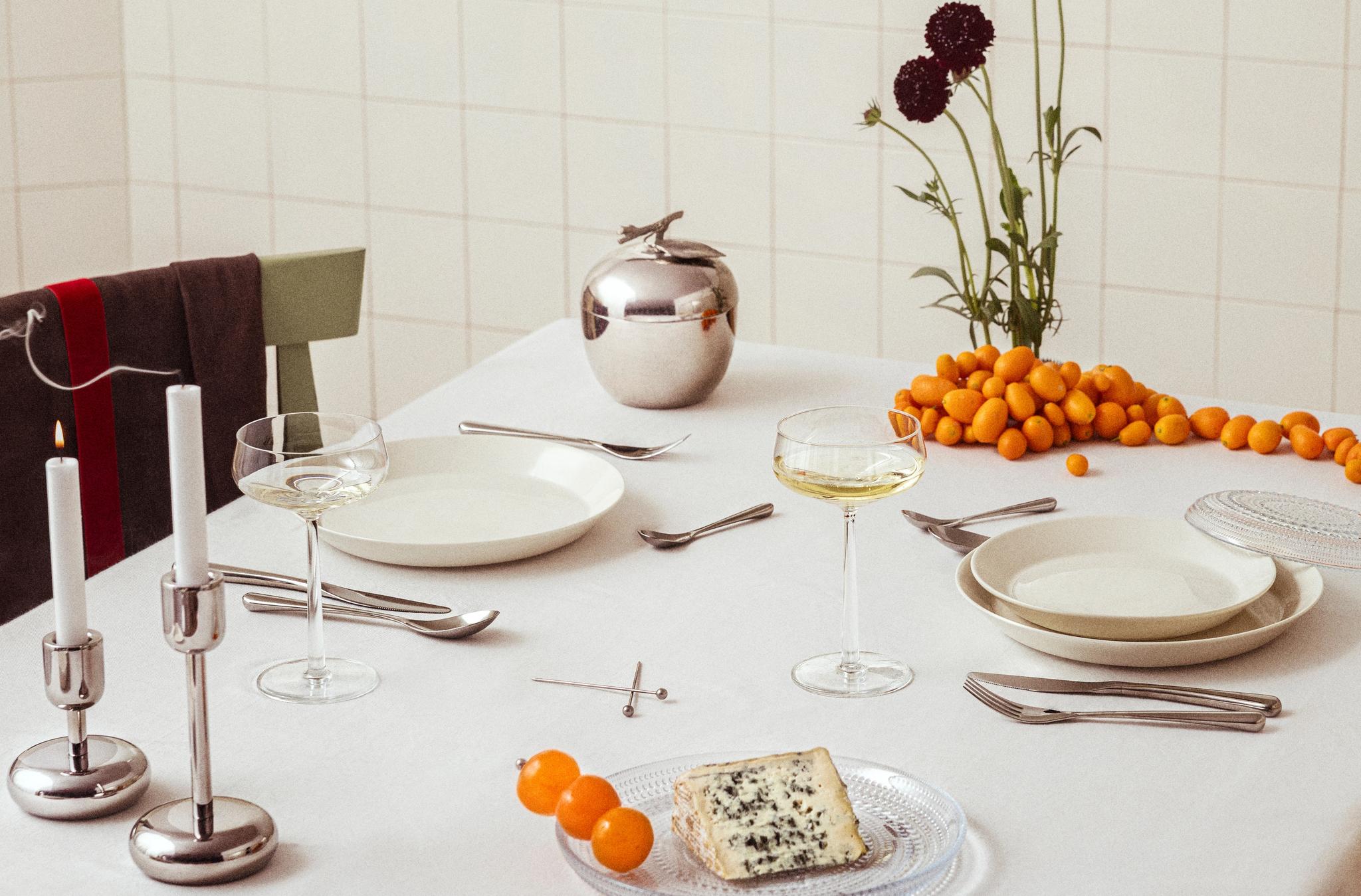 Ruokapöytä, jossa valkoiset lautaset, samppanjalasit, sytytettyjä kynttilöitä, hopeinen omenanmuotoinen rasia, tummanpunaisia kukkia, sinihomejuustoa ja kumkvatteja valkoisella pöytäliinalla.