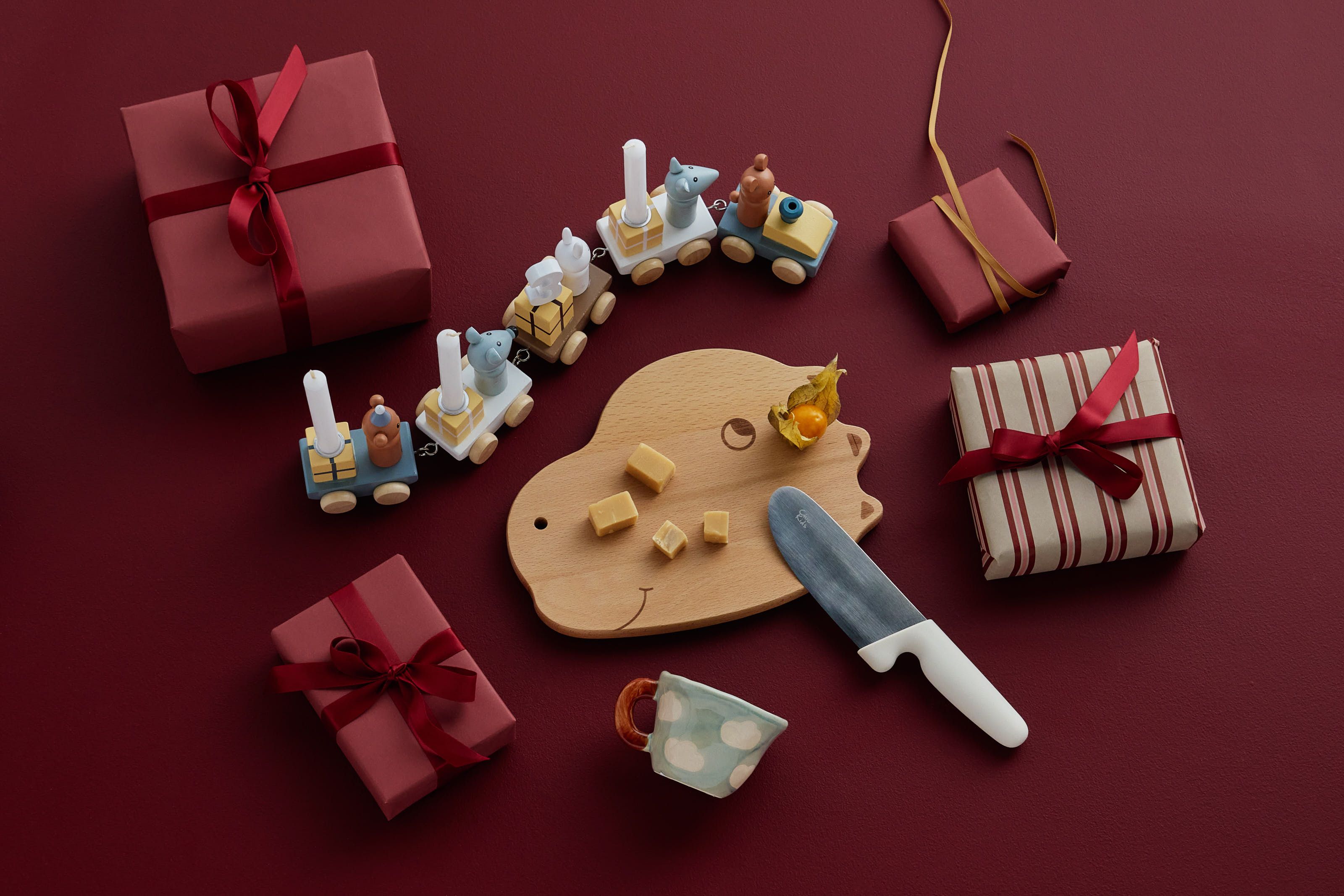 Presentinslagna askar, ett leksakståg och ett serveringsbräda av trä med ost och physalisfrukter mot röd bakgrund.