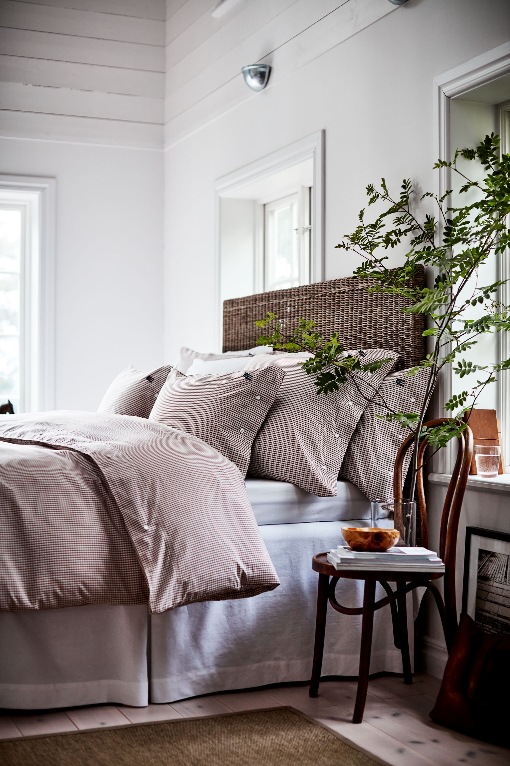 A bright bedroom with a woven wicker headboard, pink gingham bedding, a leafy plant, and a wooden side chair.