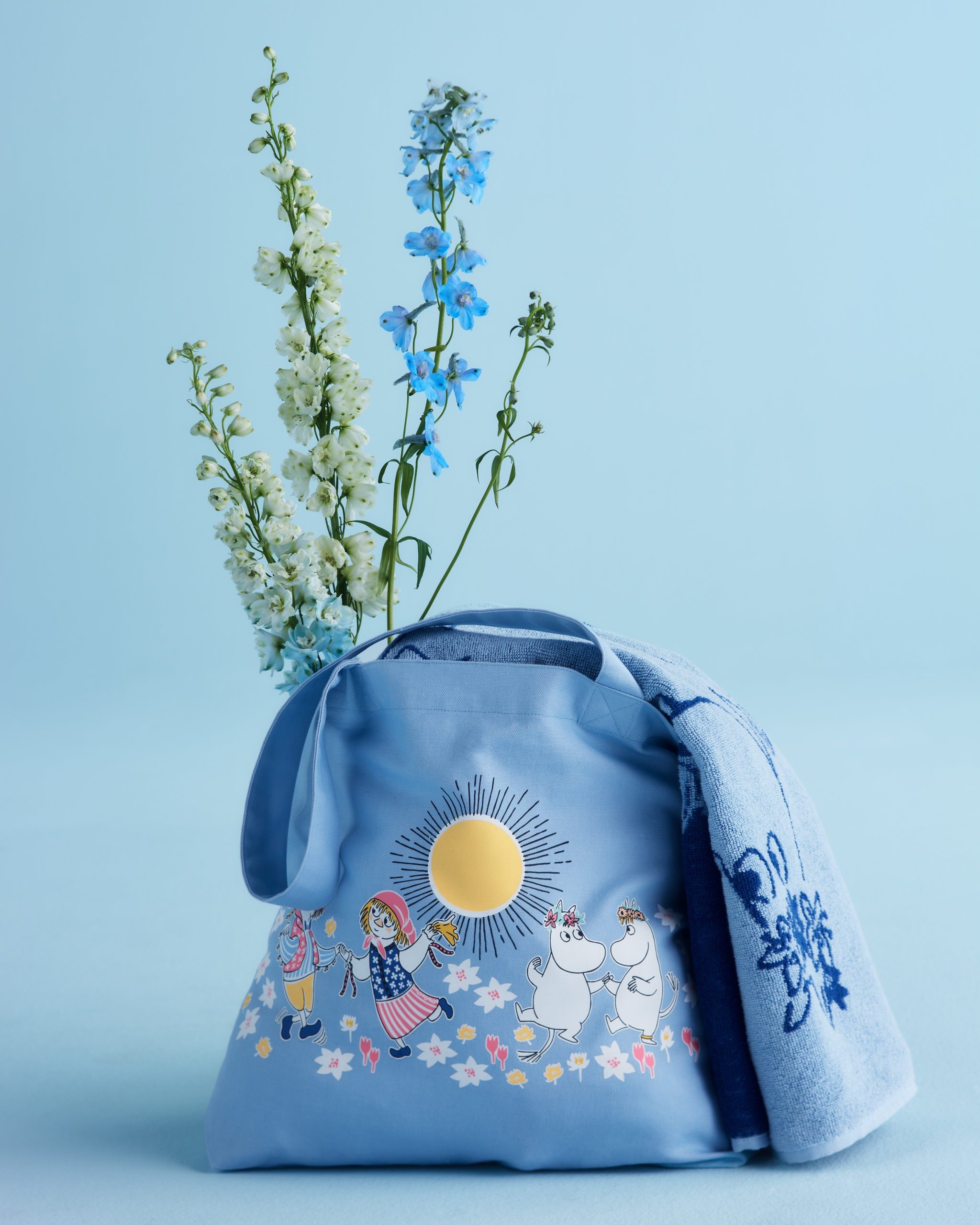 Ljusblå tygkasse med Muminmotiv, blommor och sol står mot blå bakgrund bredvid en matchande handduk och sommarblommor.