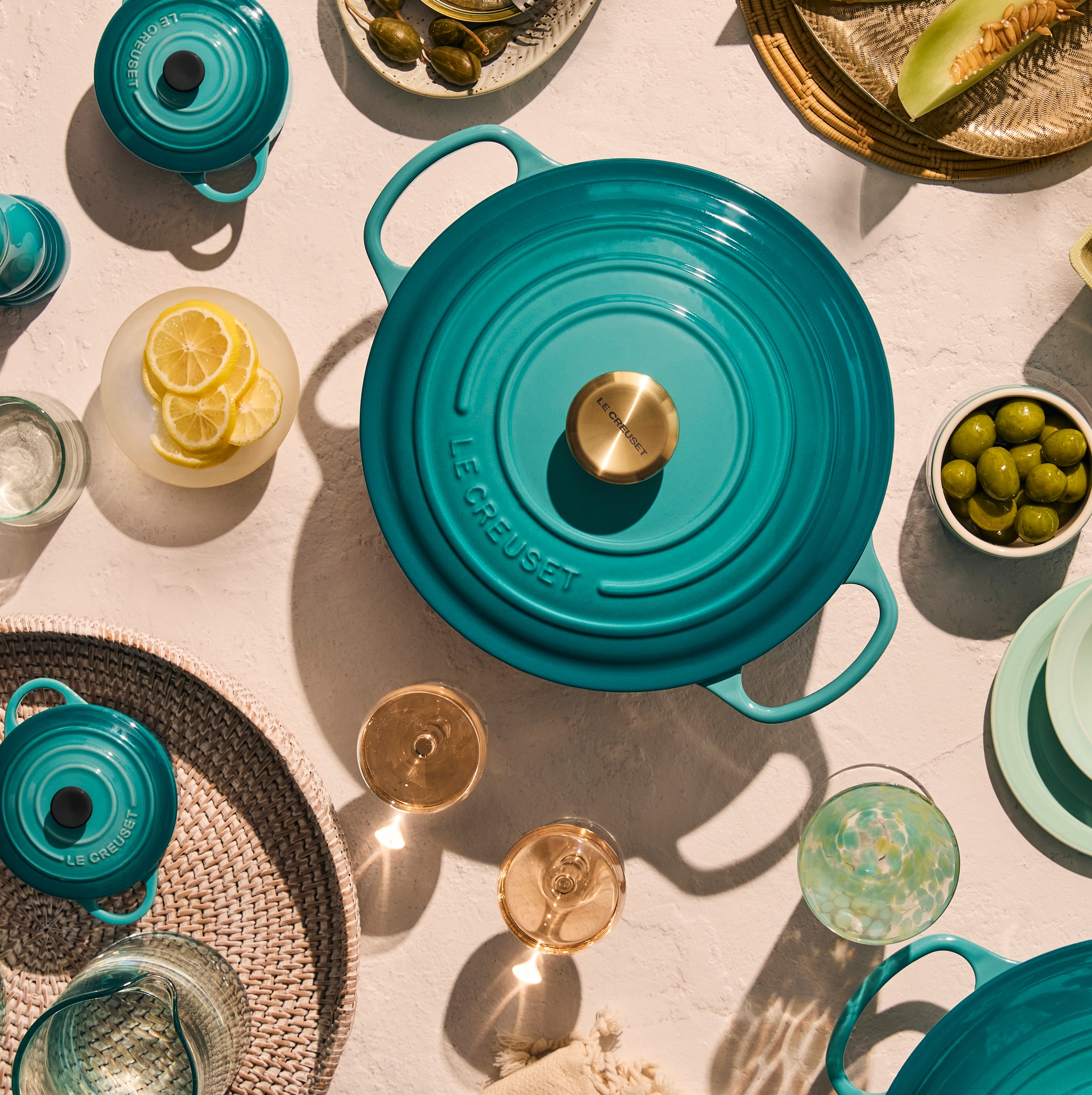 Bild ovanifrån av ett blågrönt Le Creuset-lock, två glas vitt vin och en flätad korg på en texturerad yta med skarpa skuggor.