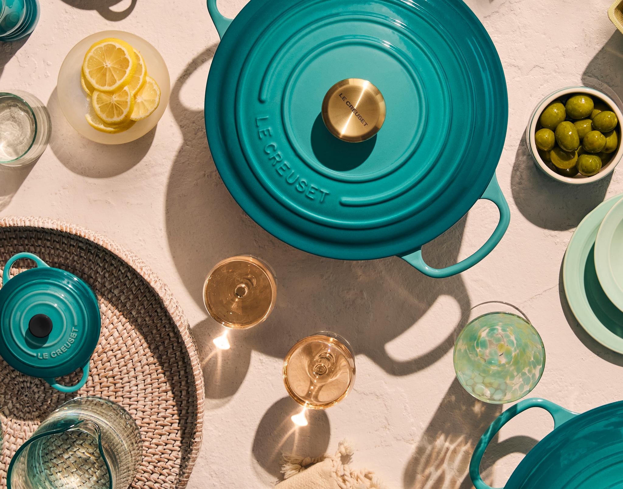 Bild ovanifrån av ett blågrönt Le Creuset-lock, två glas vitt vin och en flätad korg på en texturerad yta med skarpa skuggor.