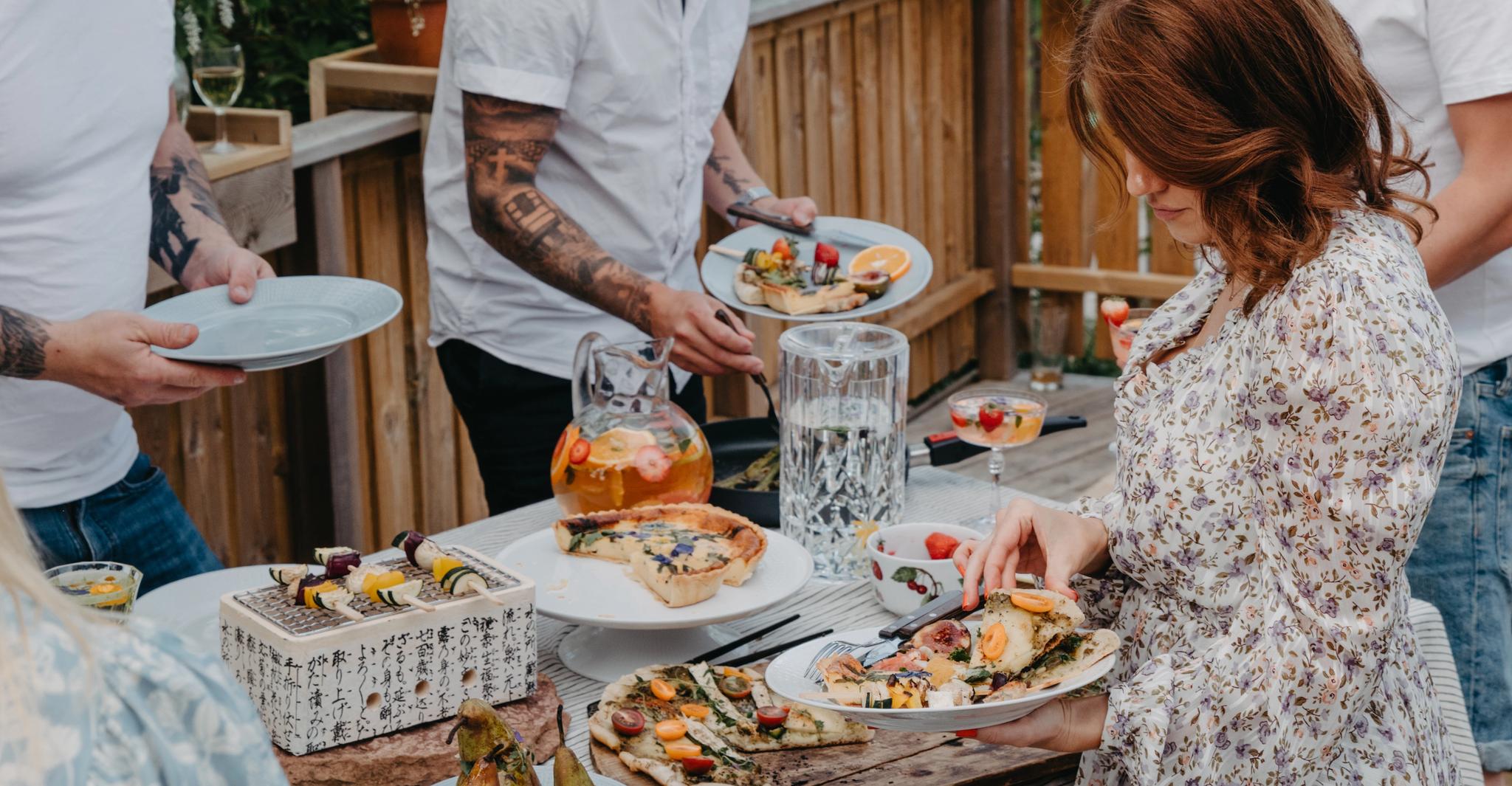 Vänner samlas runt ett dukat bord på en solig altan och serverar sig mat under en somrig utomhusmiddag.