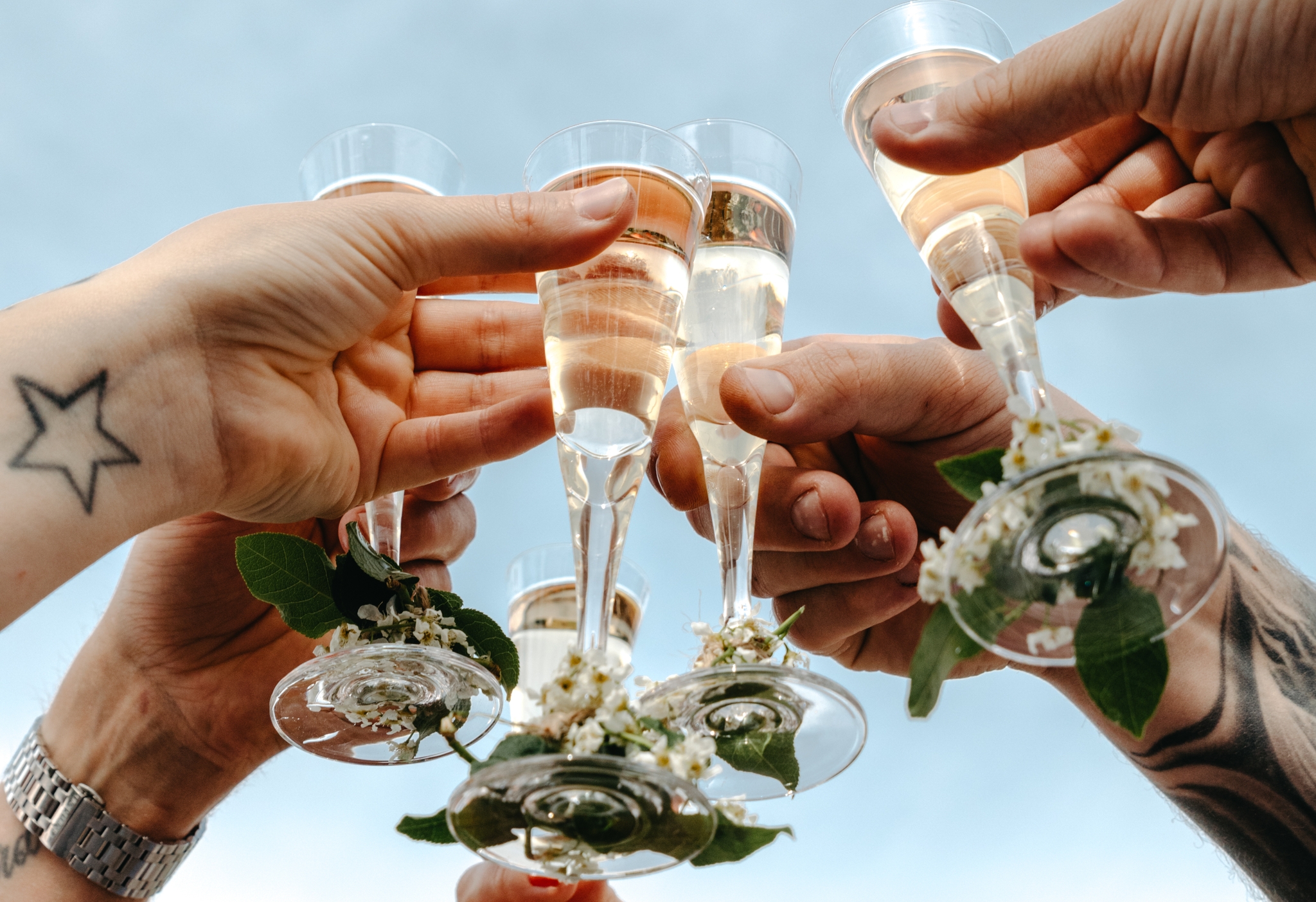 Flera händer skålar med bubbel i champagneglas dekorerade med blommor mot en blå himmel.