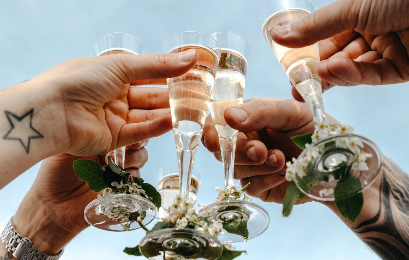 Flere hender skåler med champagneglass pyntet med blomster mot en blå himmel.