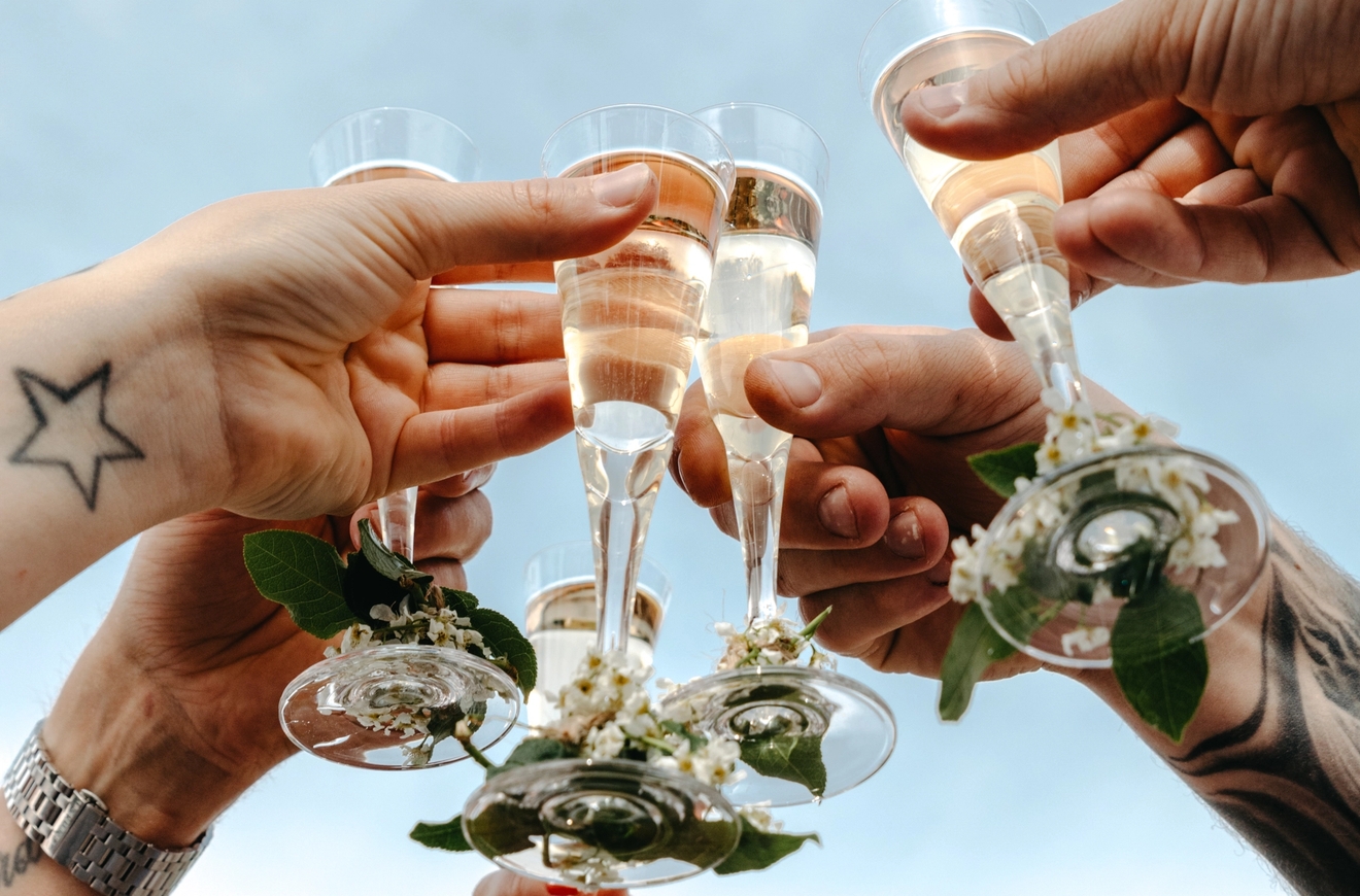 Flere hender skåler med champagneglass pyntet med blomster mot en blå himmel.