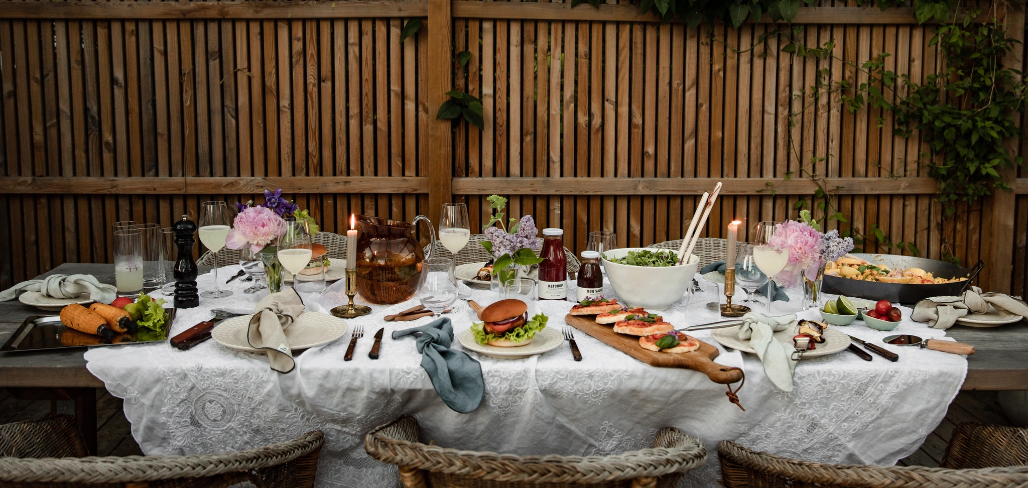 Ett utomhusdukad middagsbord fyllt med mat, dryck och blommor, upplyst av ljusslingor