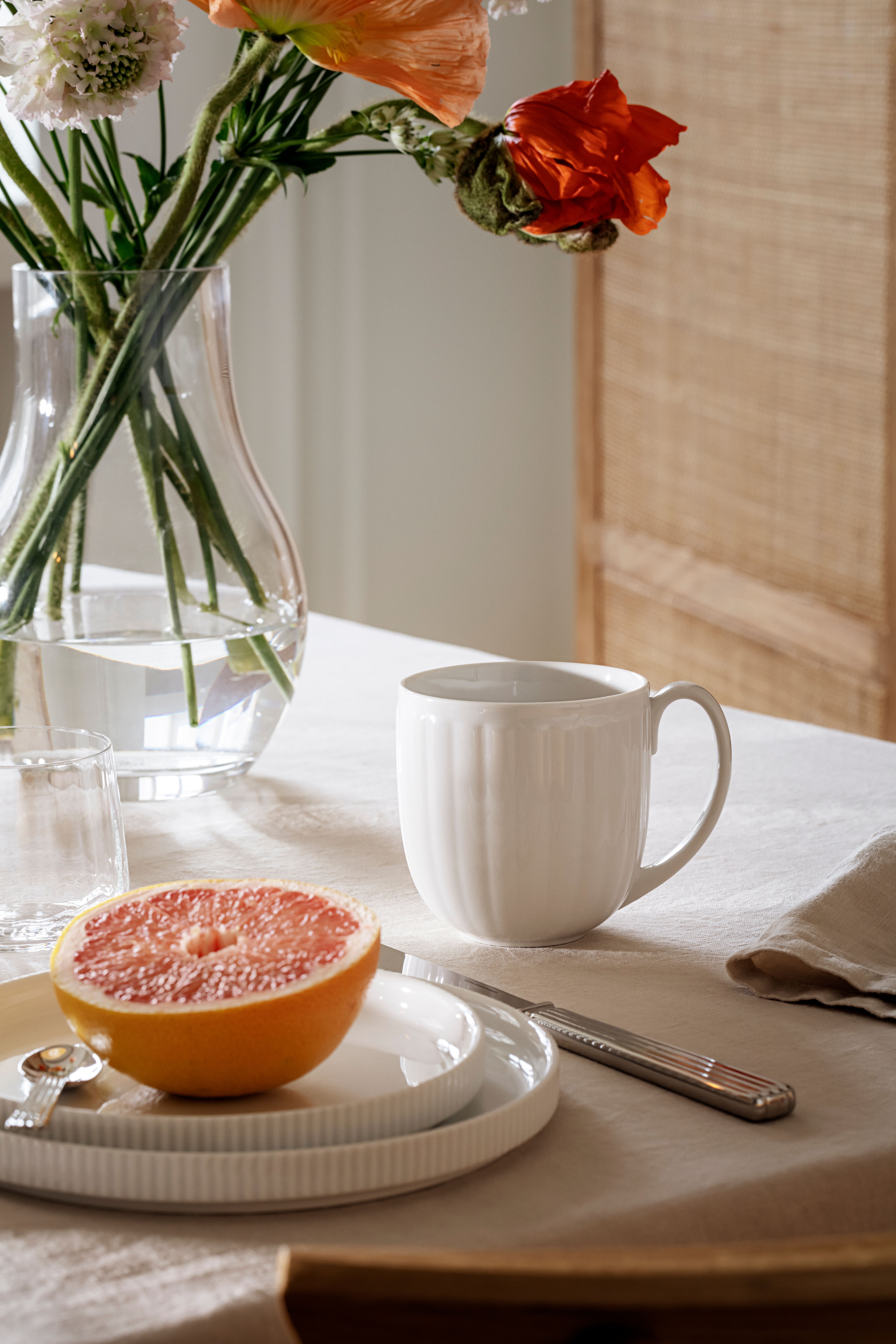 Ett bord med en vit mugg, skivad grapefrukt på tallrikar och en vas med apelsin- och vita blommor.