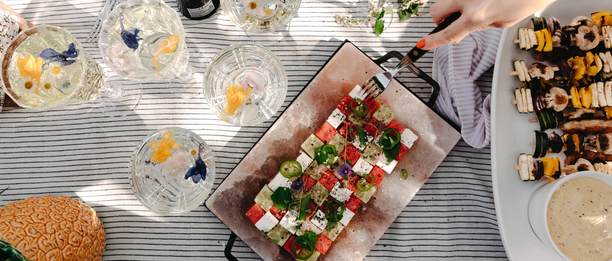 Sommerlig piknikbord ovenfra med fargerik salat, grillspyd, drinker med blomster og musserende vin.