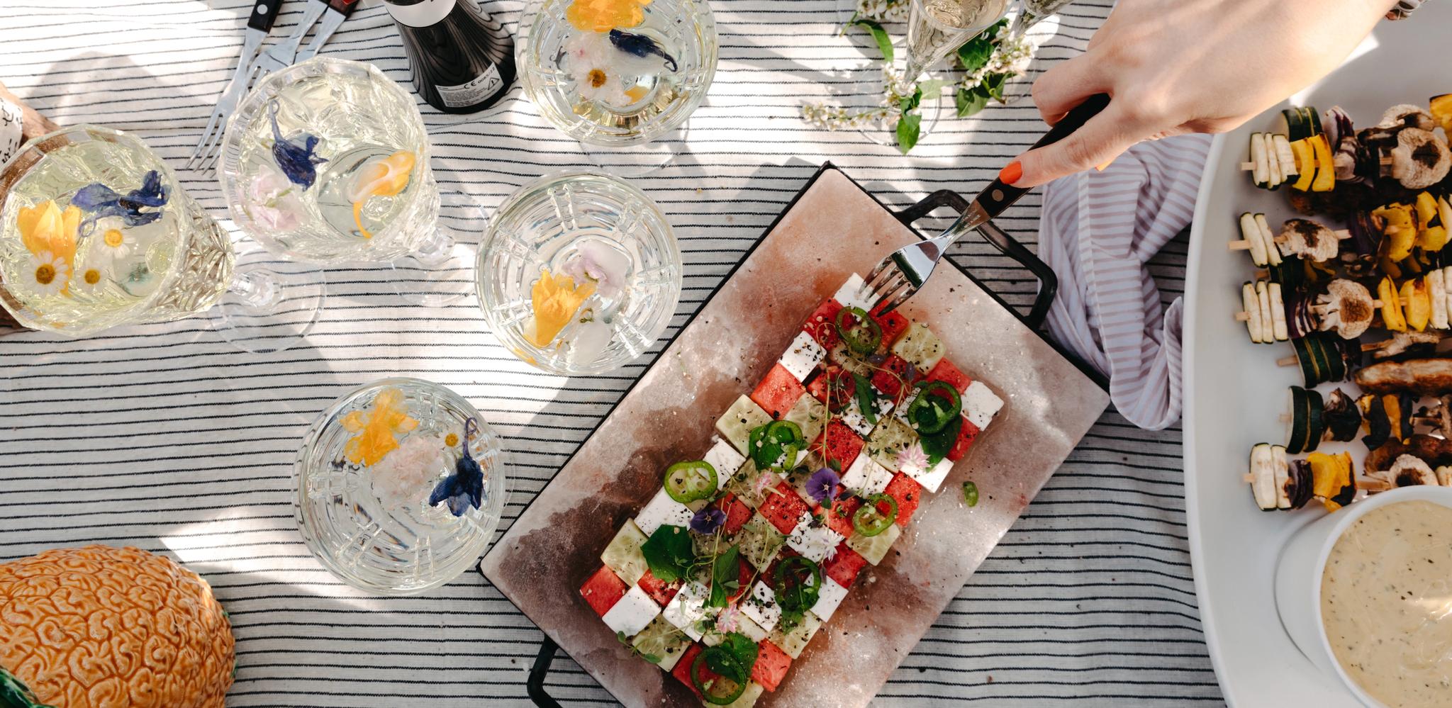 Sommerlig piknikbord med fargerik salat, grillspyd, bobler og drinker pyntet med spiselige blomster.