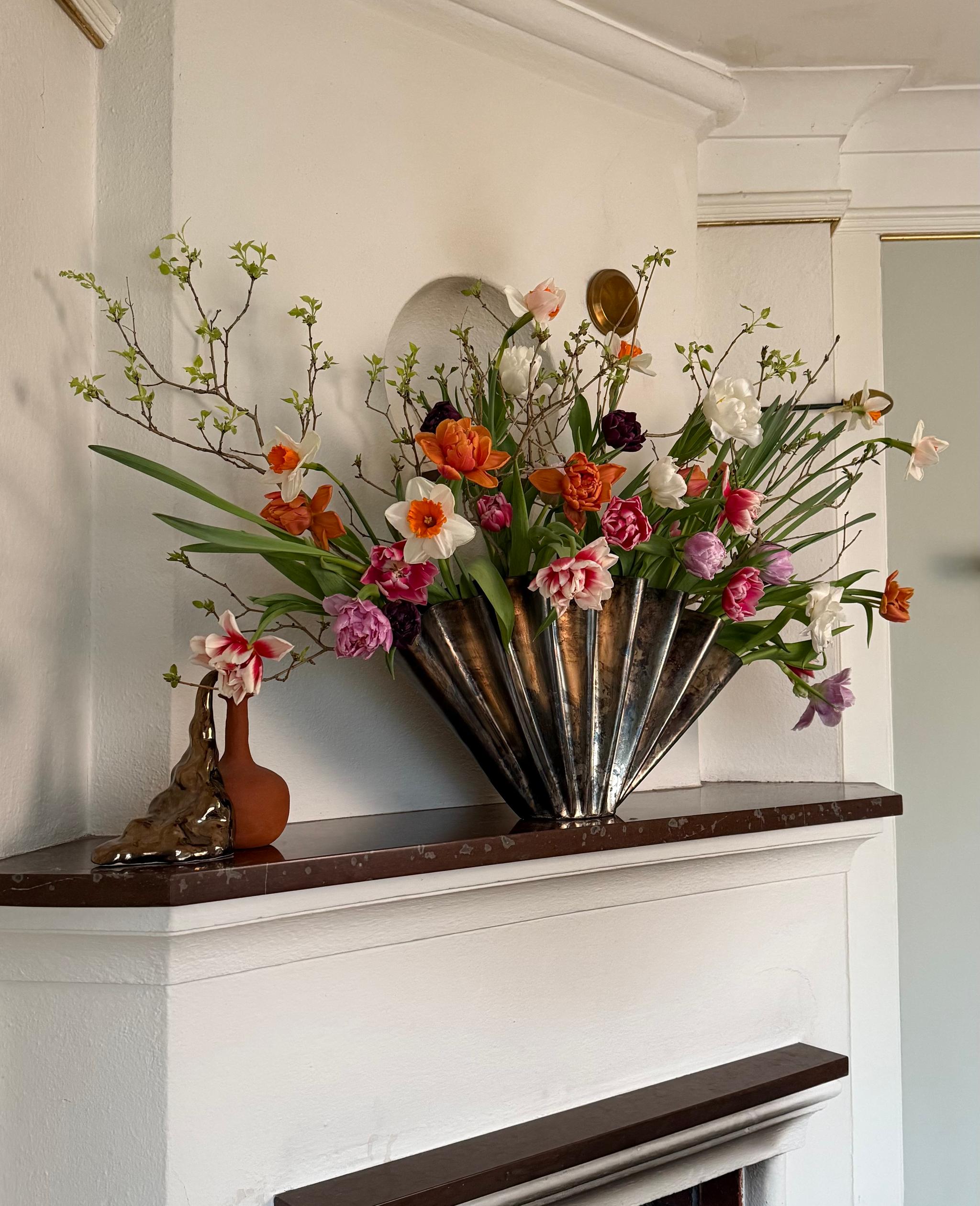 A large, colorful spring flower arrangement with tulips and daffodils in a metallic fan-shaped vase on a white fireplace mantel.