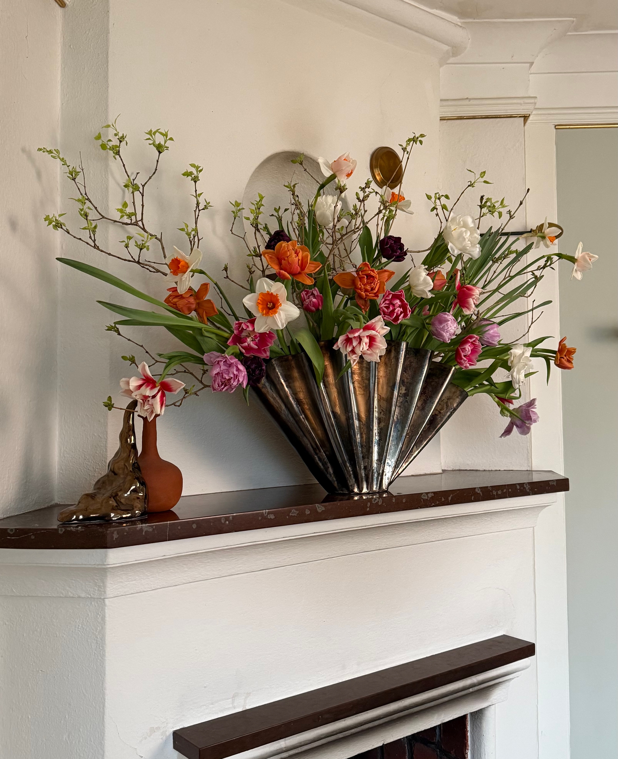 A vibrant arrangement of orange, pink, and white tulips and daffodils in a fanned metallic vase on a white fireplace mantel.
