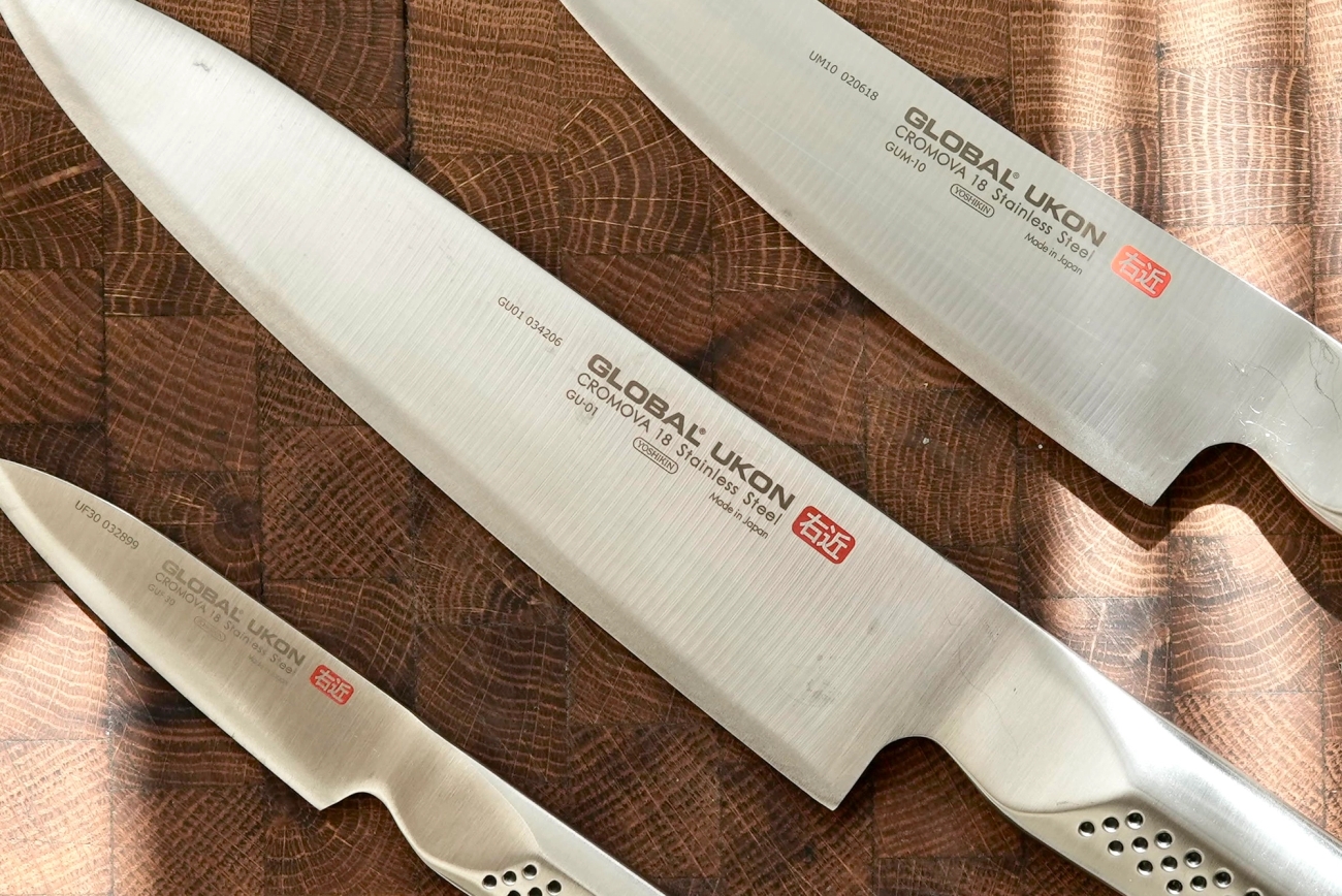 Tre japanska Global Ukon-kockknivar i rostfritt stål ligger på en skärbräda av ändträ i trä.