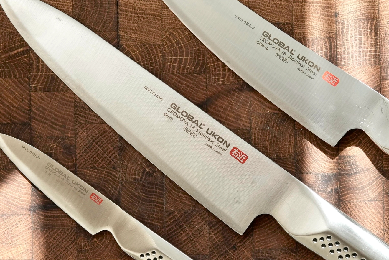 Tre japanska Global Ukon-kockknivar i rostfritt stål ligger på en träskärbräda.