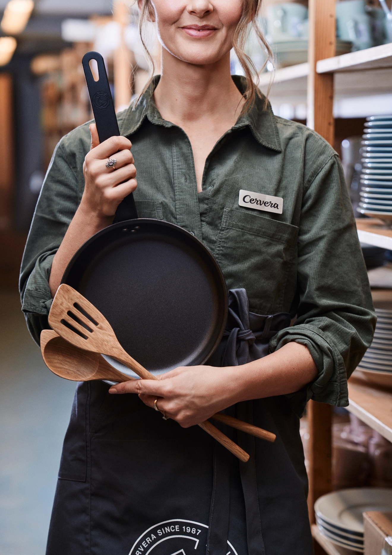 Nainen Cervera-esiliina yllään, kädessään musta paistinpannu ja puiset lastat