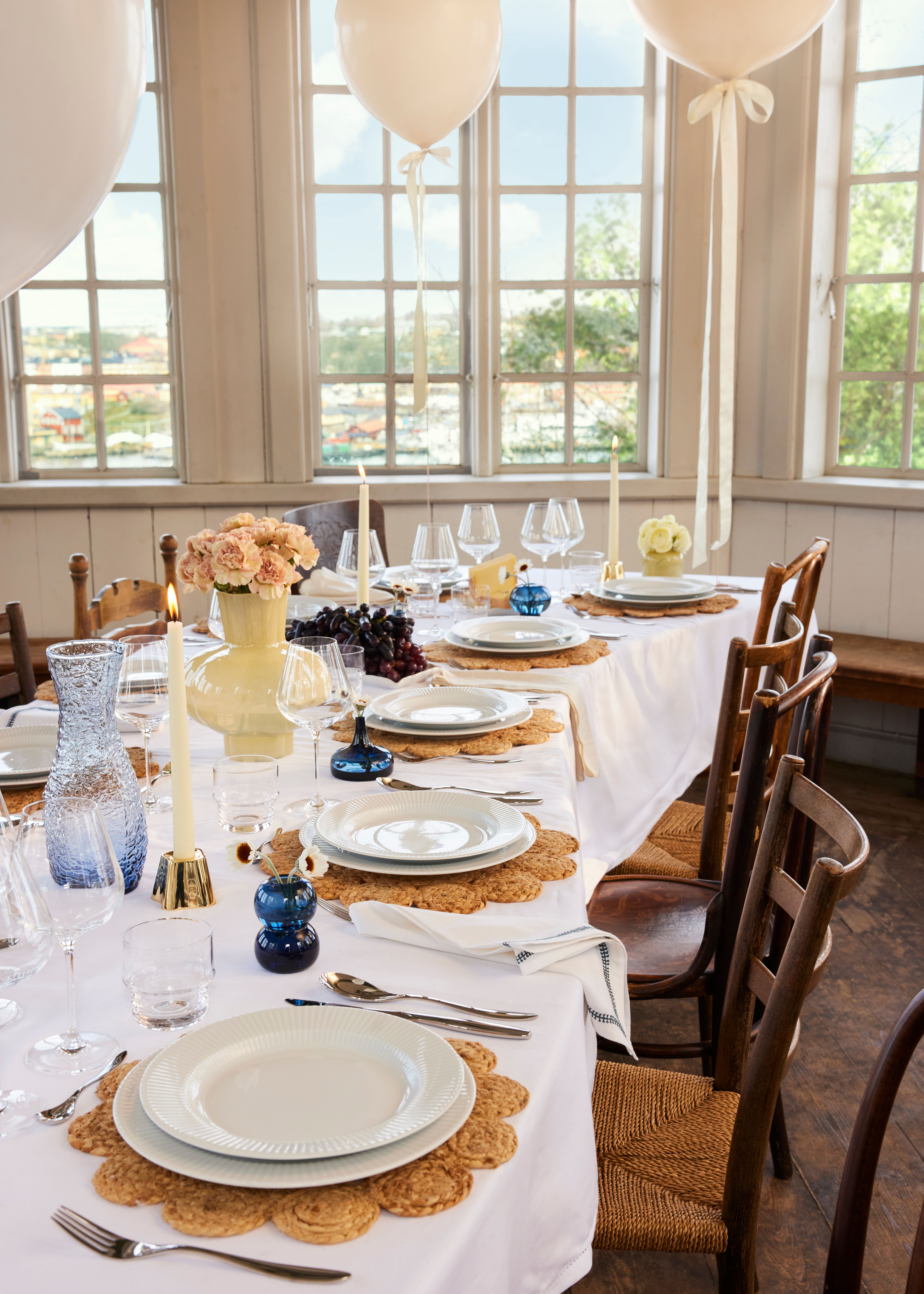 An elegantly set long dining table with white plates on woven placemats, candles, flowers, and white balloons, bathed in light from large windows.