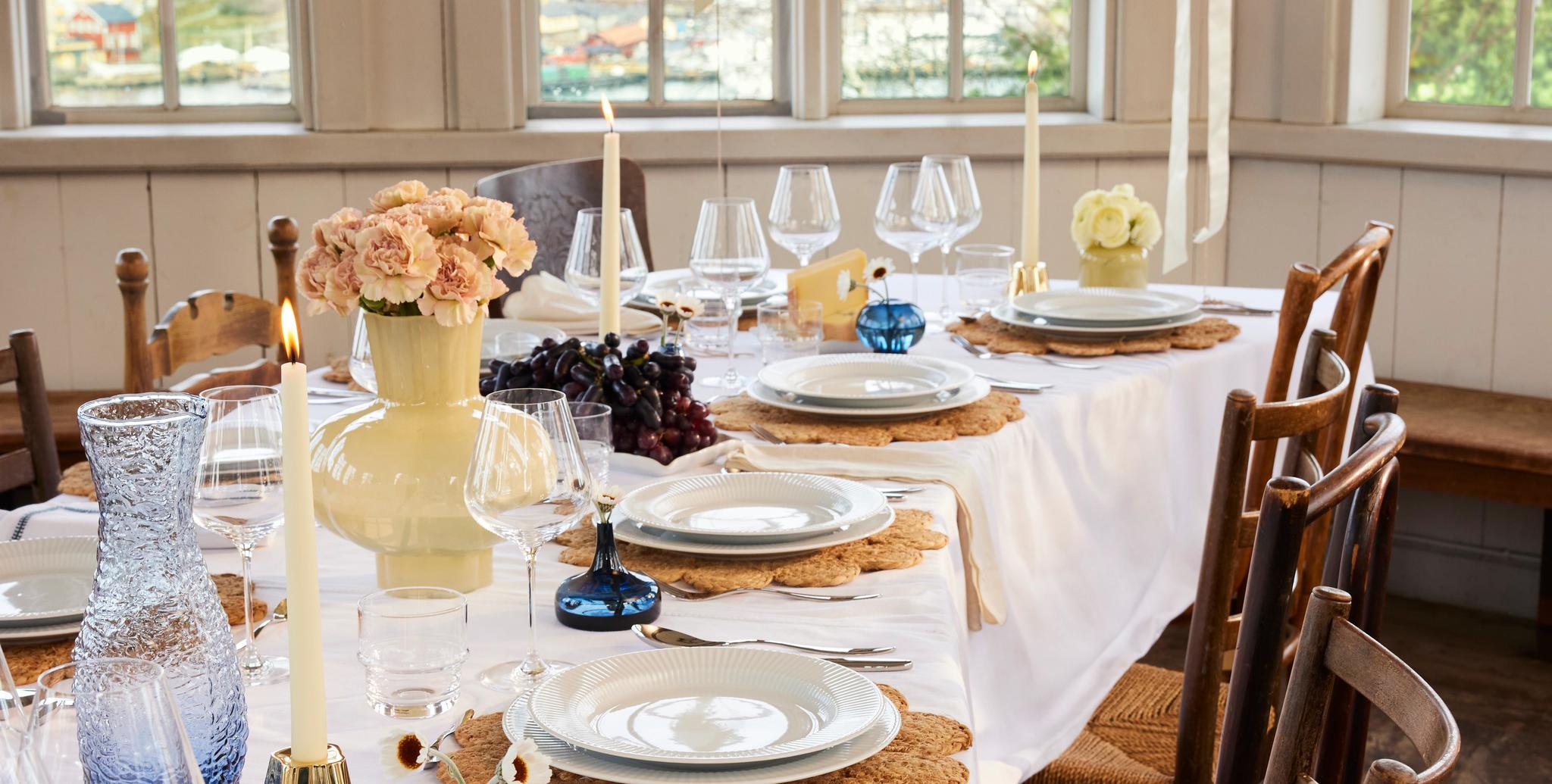 An elegantly set long dining table with white plates on woven placemats, candles, flowers, and white balloons, bathed in light from large windows.