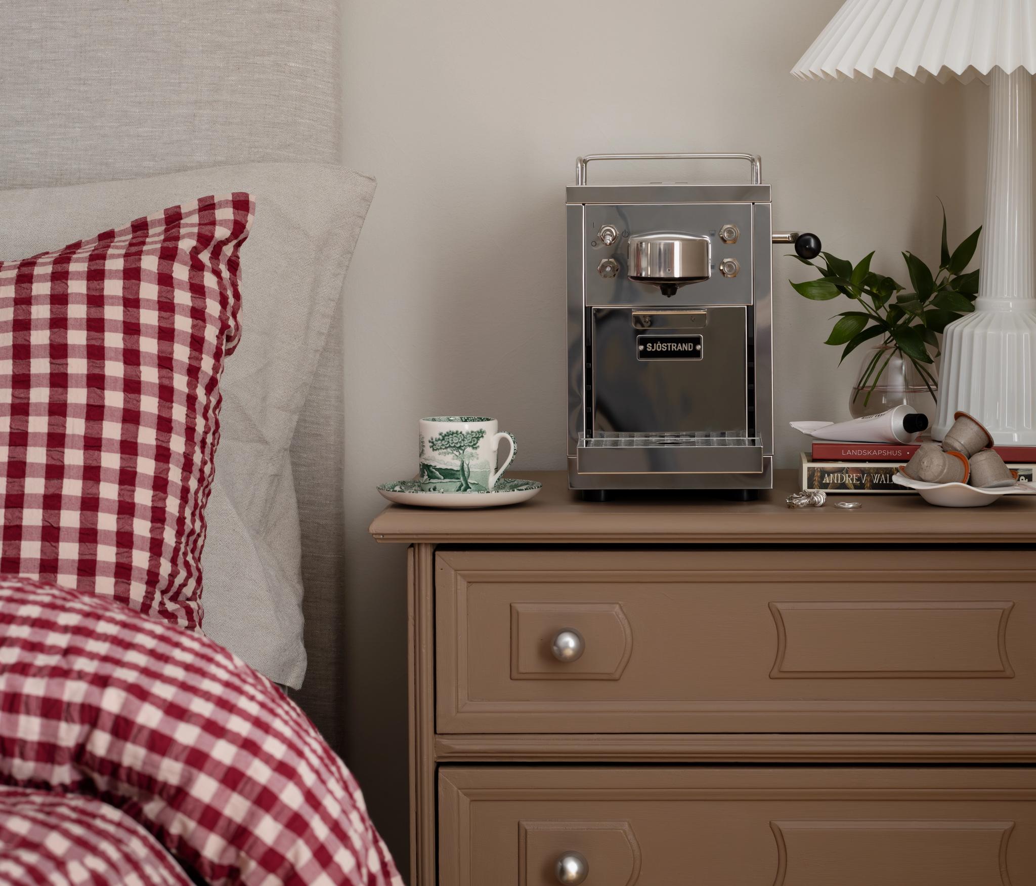 A silver espresso machine, teacup, and white pleated lamp on a brown nightstand next to a bed with red gingham bedding.