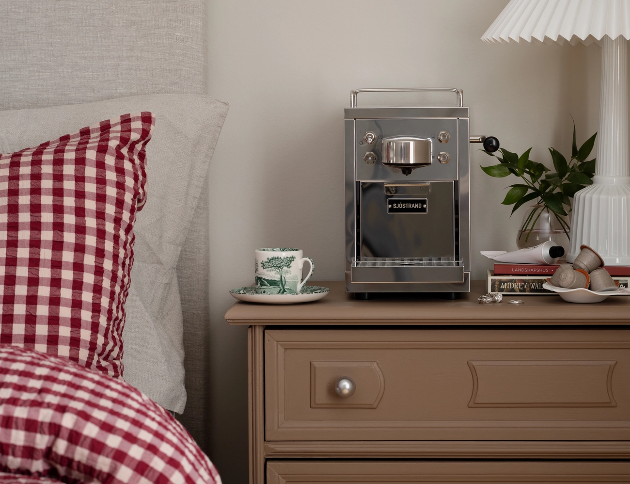 A brown bedside table holds a silver espresso machine, a patterned teacup, and a white pleated lamp, next to a bed with red gingham bedding.