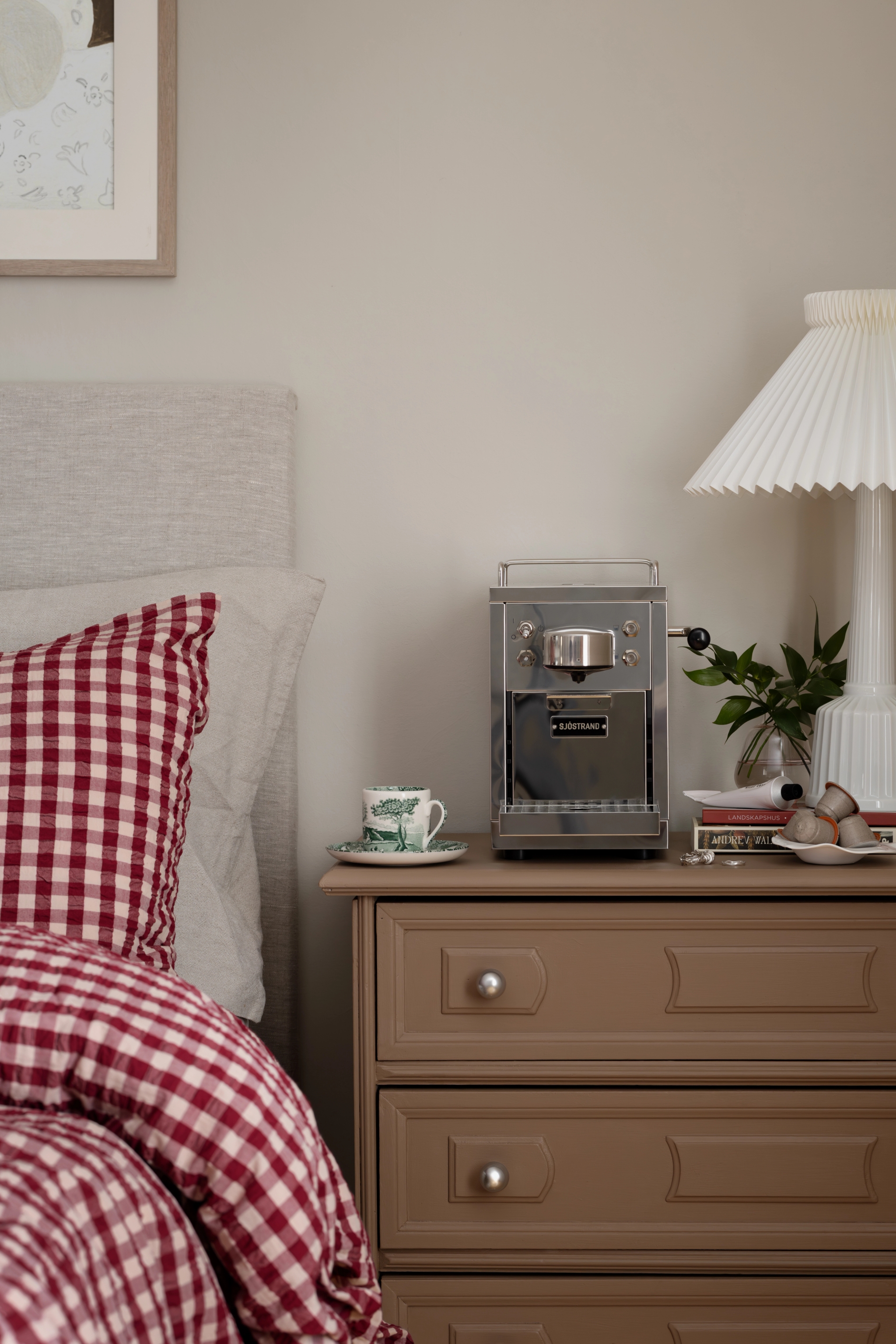 A silver espresso machine, teacup, and white pleated lamp on a brown nightstand next to a bed with red gingham bedding.