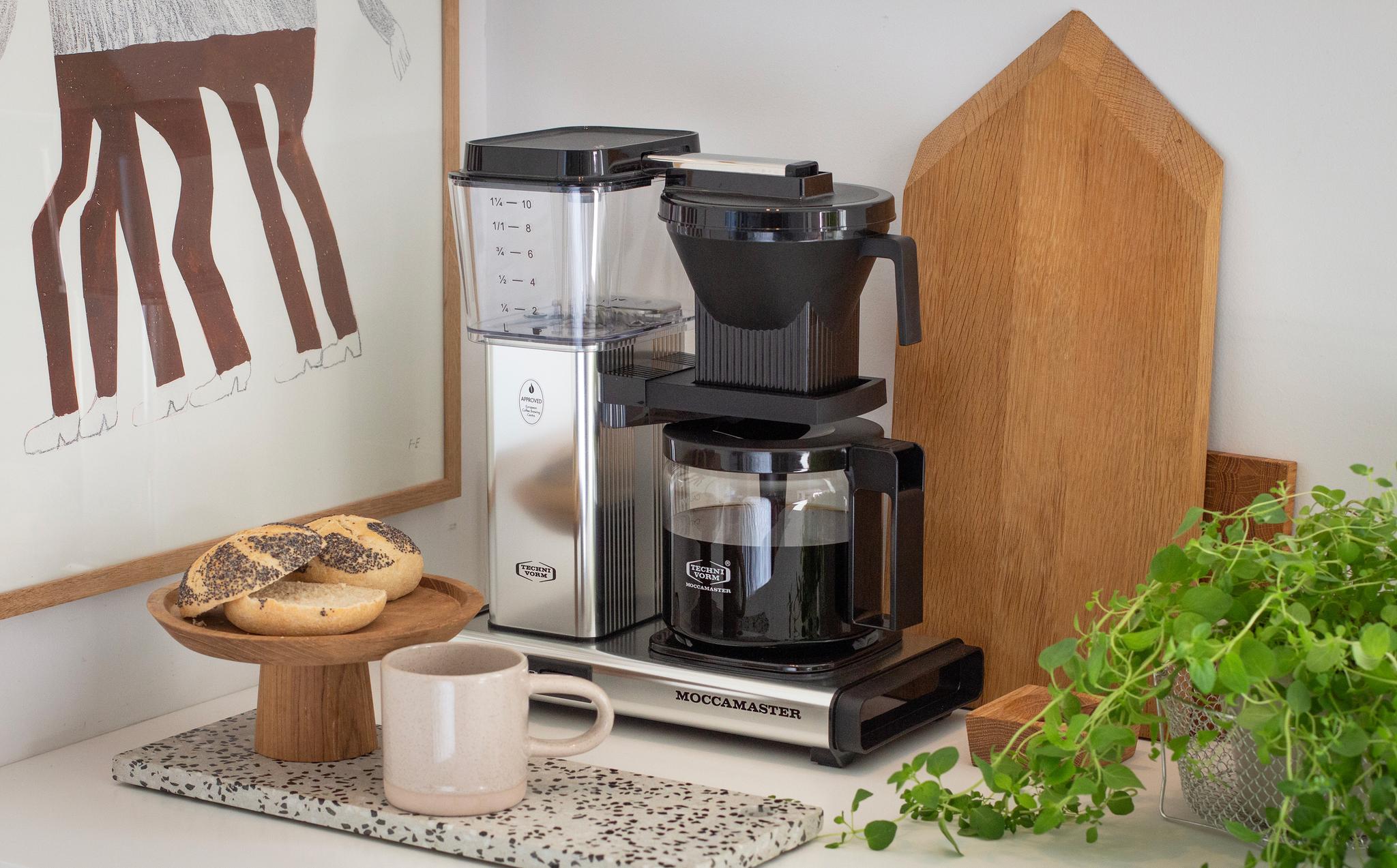 Moccamaster kaffebryggare på köksbänk med nybryggt kaffe, frallor, mugg och gröna växter.