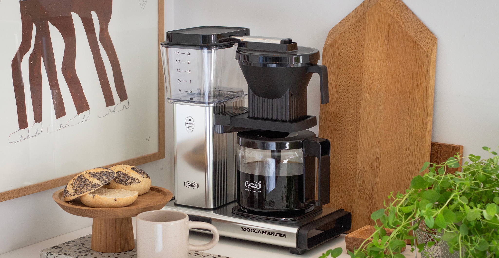 Stilren Moccamaster kaffetrakter på kjøkkenbenk med nytraktet kaffe, rundstykker og grønn plante.
