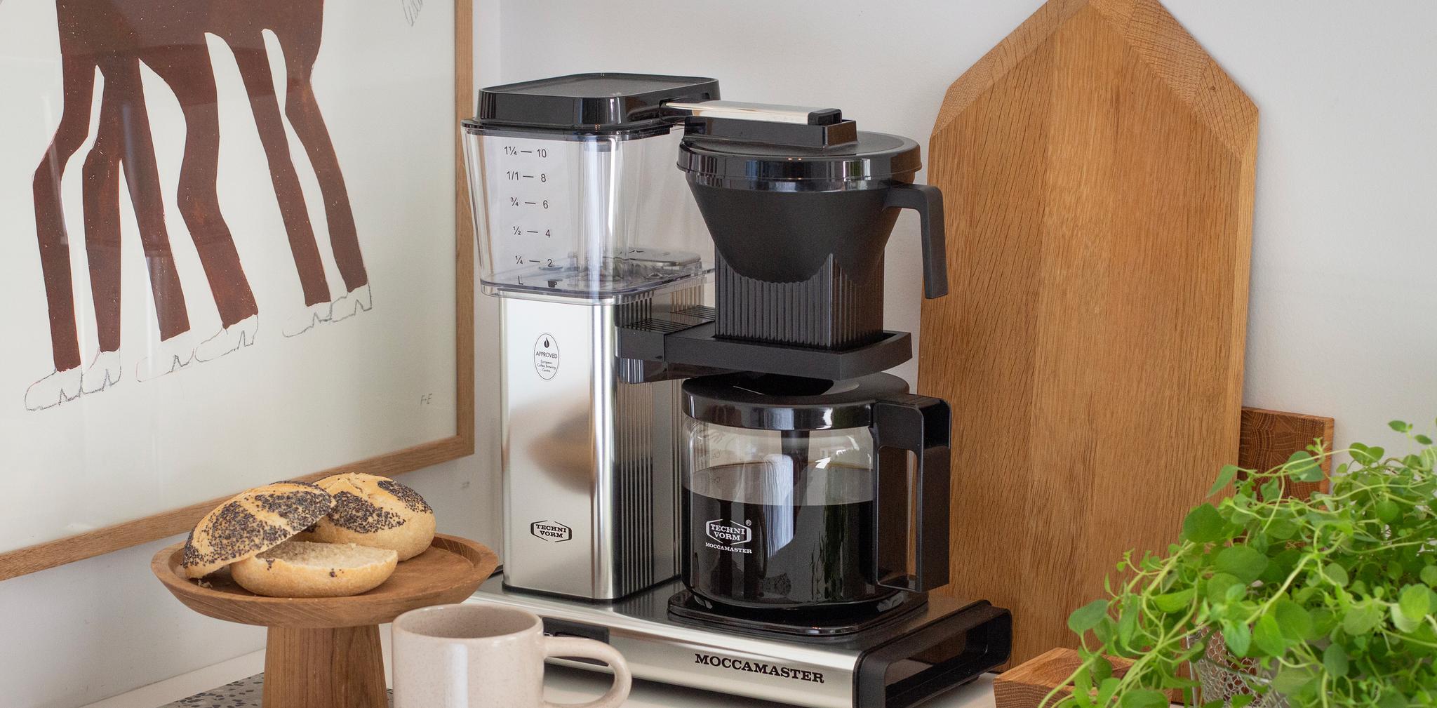Moccamaster kaffetrakter på kjøkkenbenk med nytraktet kaffe, rundstykker og grønn plante.
