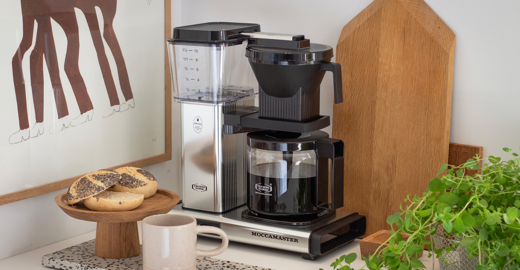 En Moccamaster kaffebryggare står på en stilren köksbänk bredvid en kopp kaffe, frallor och gröna växter.