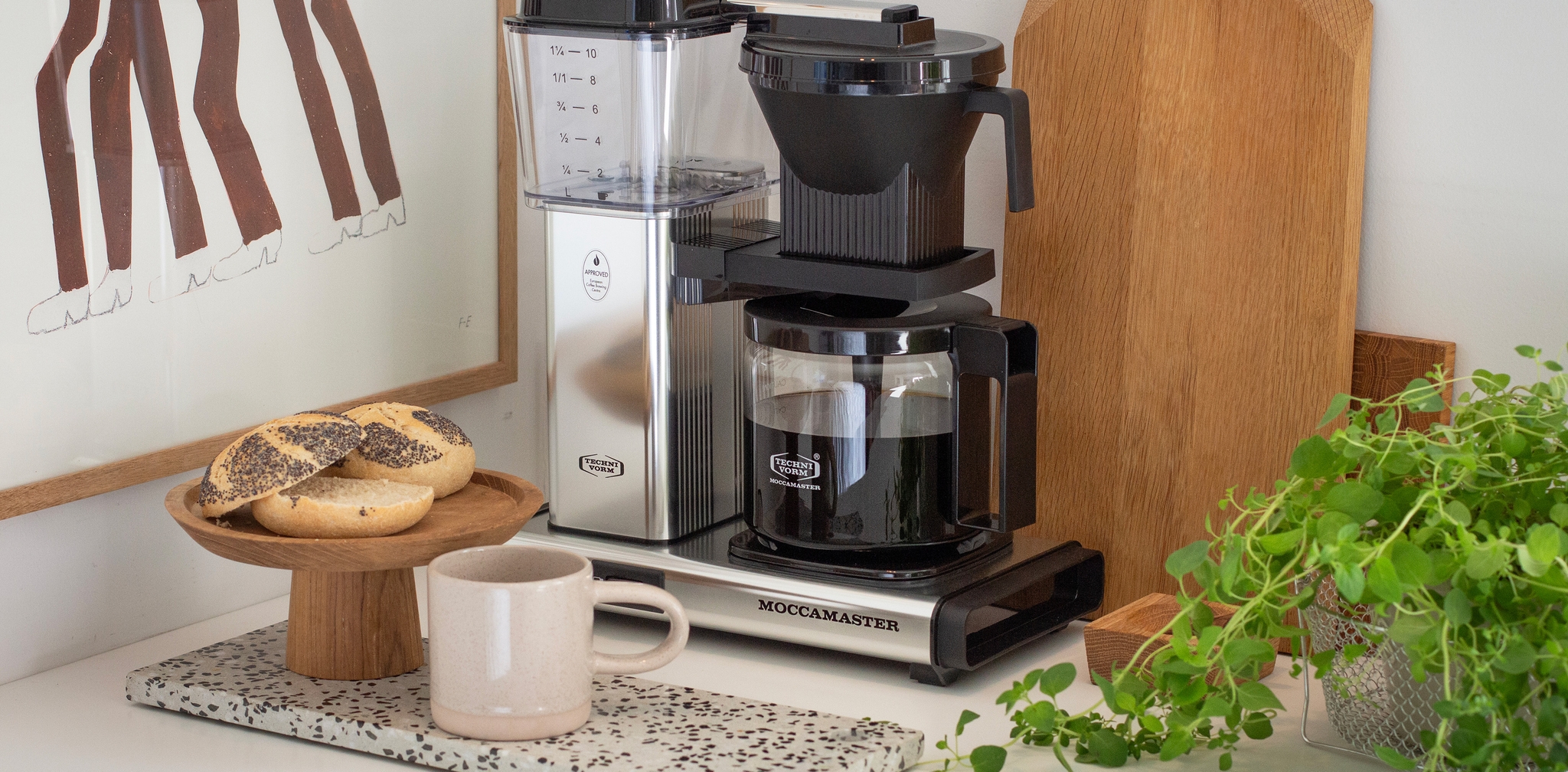 Moccamaster kaffetrakter på kjøkkenbenk med nytraktet kaffe, rundstykker og grønn plante ved siden av.