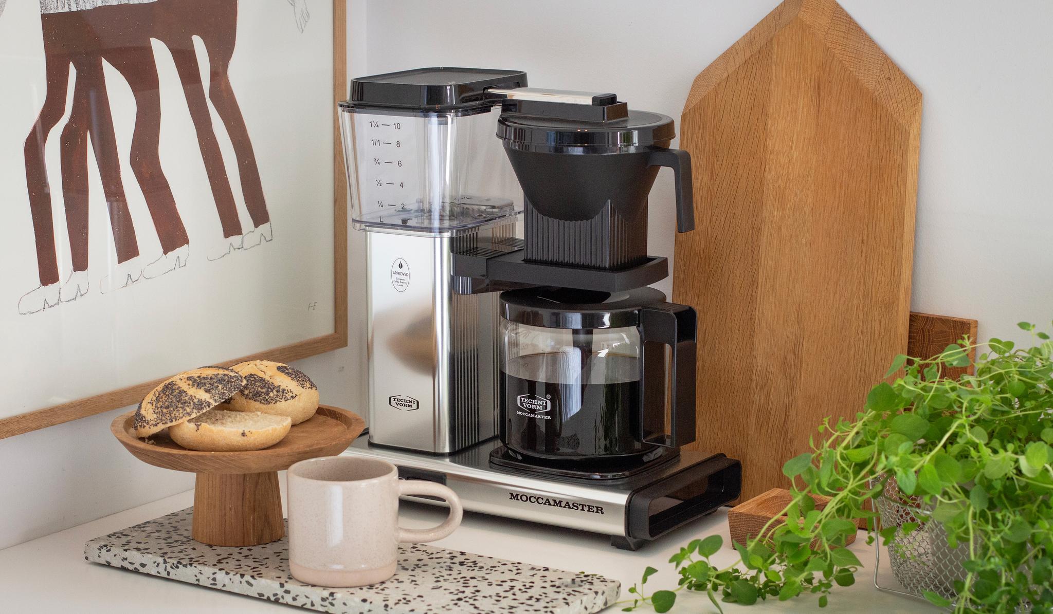 En Moccamaster kaffebryggare står på en stilren köksbänk bredvid en kopp kaffe, frallor och gröna växter.