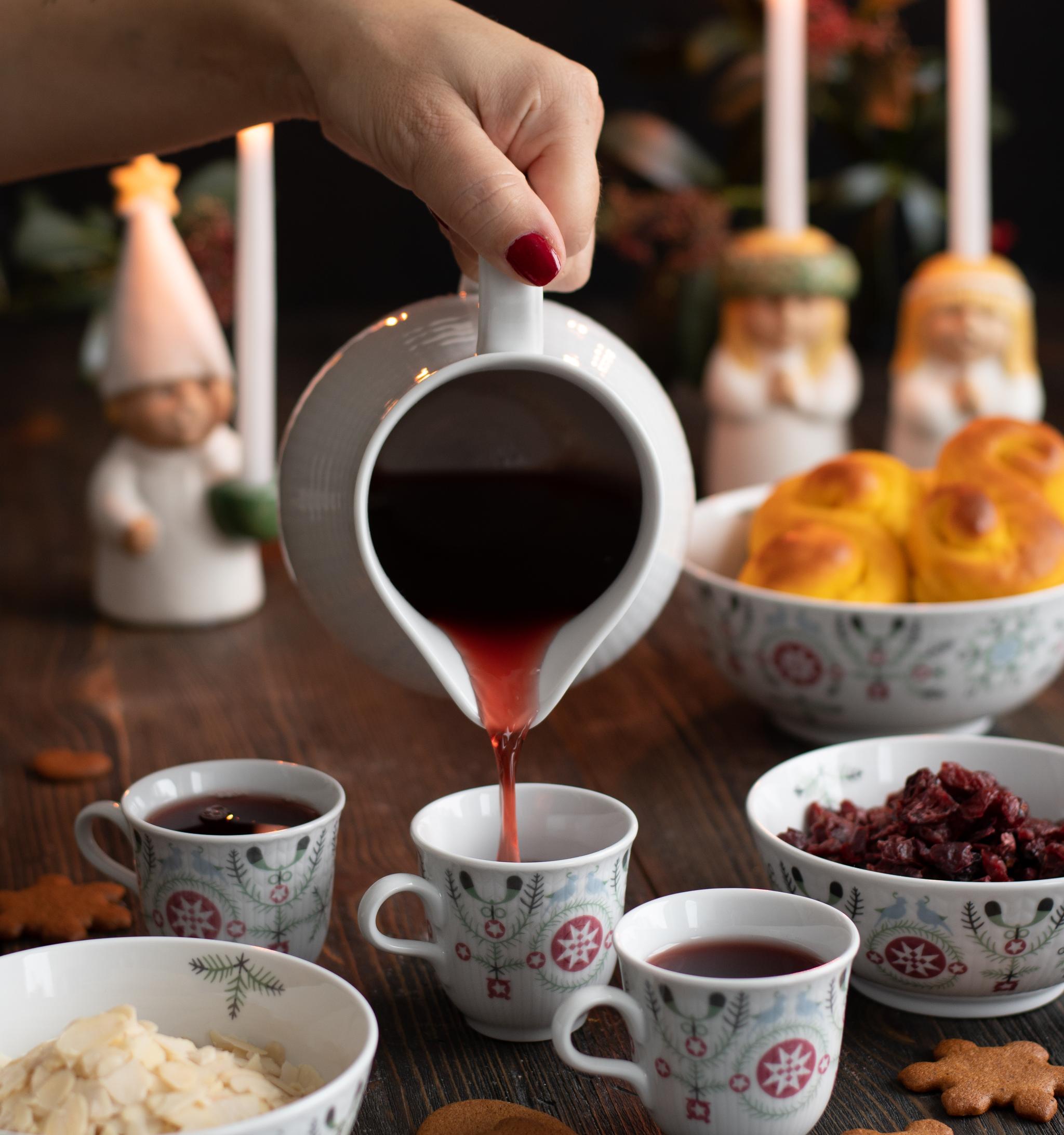 A hand pours glögg into a festive cup on a wooden table set with saffron buns, gingerbread, almonds, dried cranberries, and lit candles with gnomes.