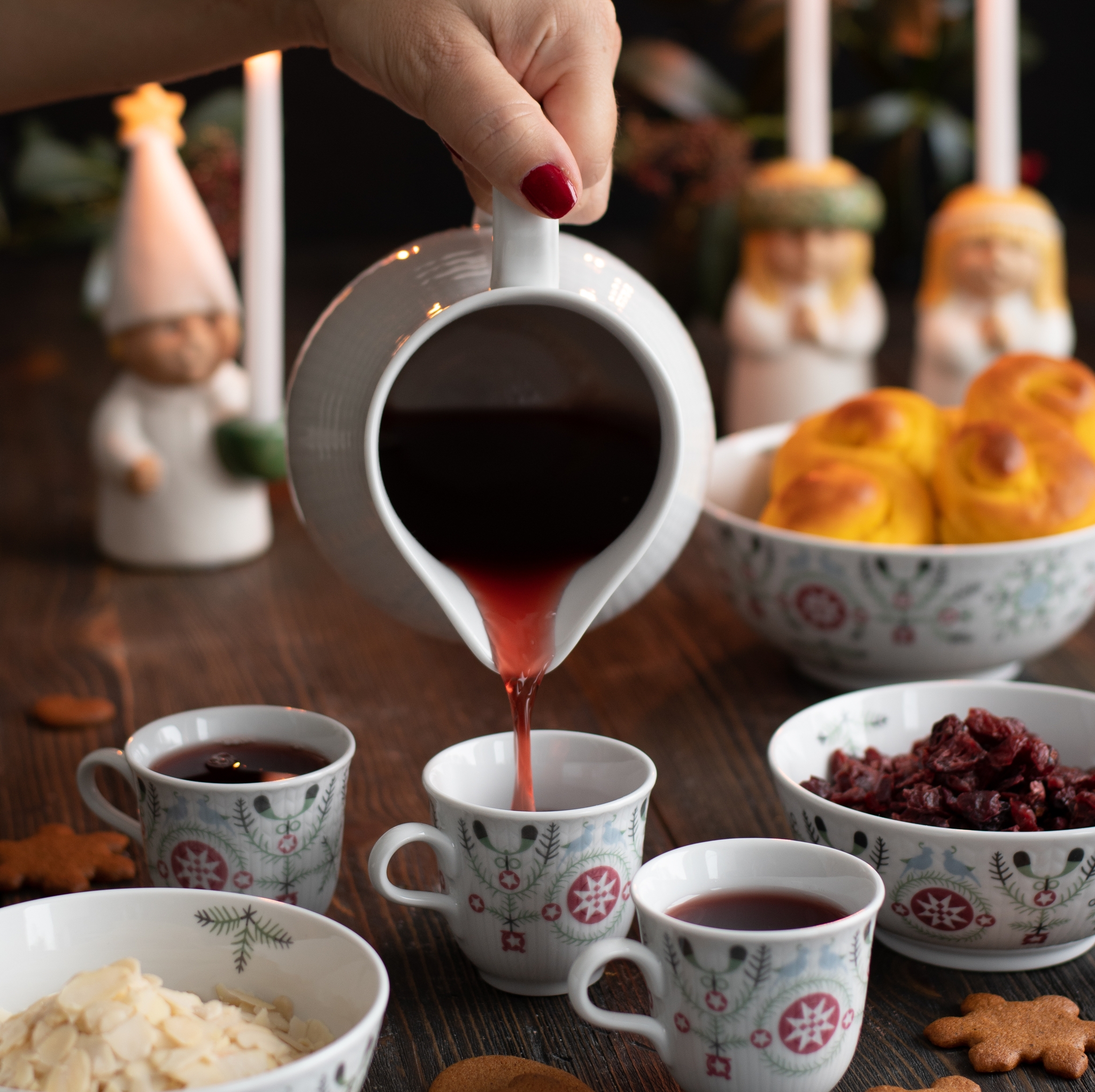 A hand pours mulled wine from a pitcher into a festive mug, surrounded by saffron buns, gingerbread cookies, toppings, and lit candles.