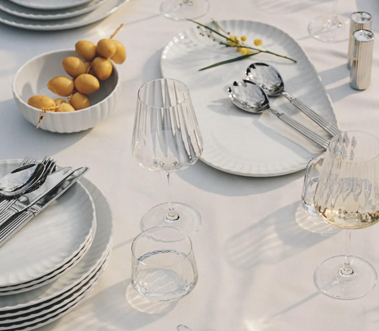 A white table set with elegant dinnerware, textured glasses, silverware, a bowl of dates, and mimosa flowers.