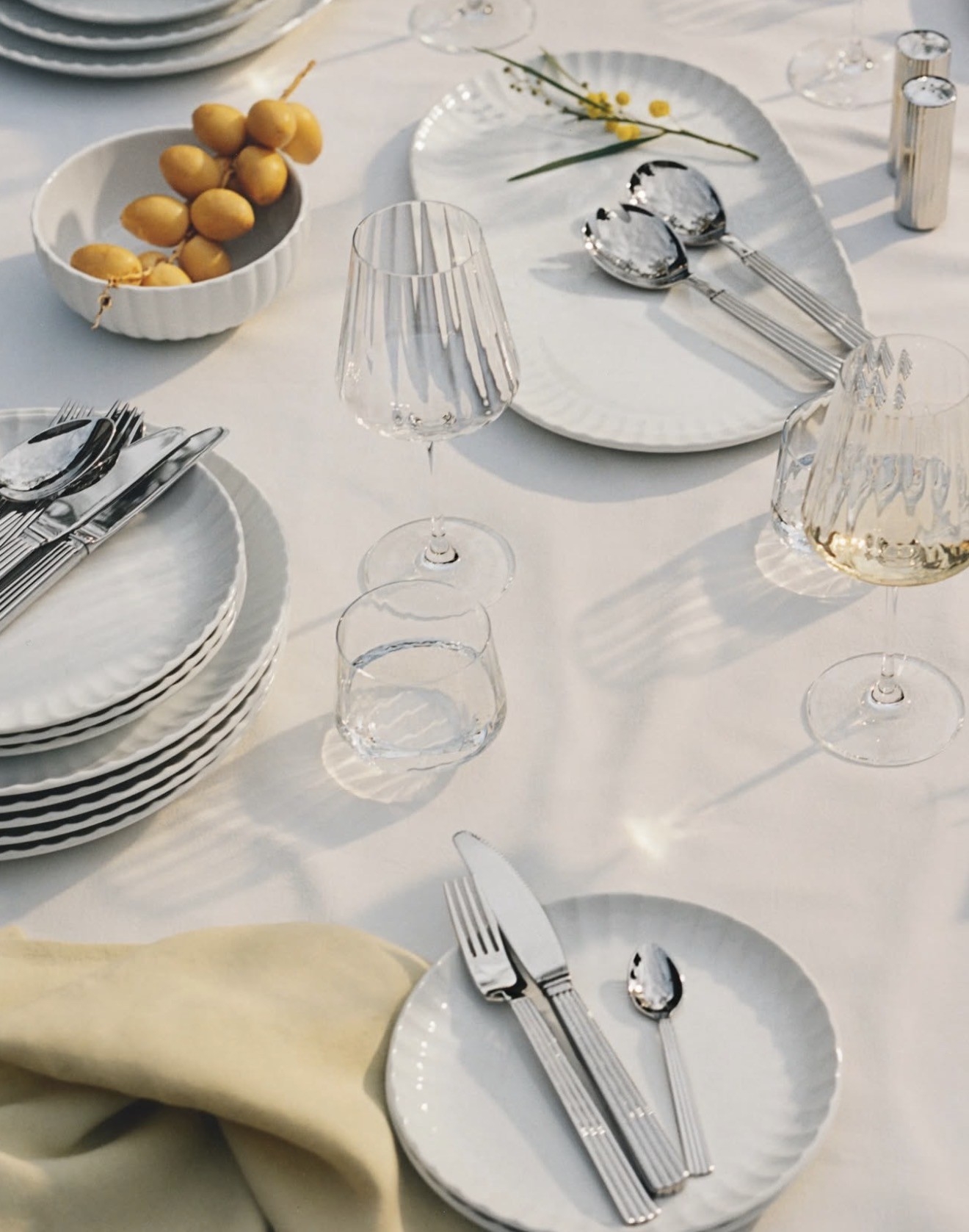 An elegant table setting featuring white ribbed plates, ribbed glassware, and silverware on a white tablecloth, with a bowl of dates and a sprig of mimosa flowers.