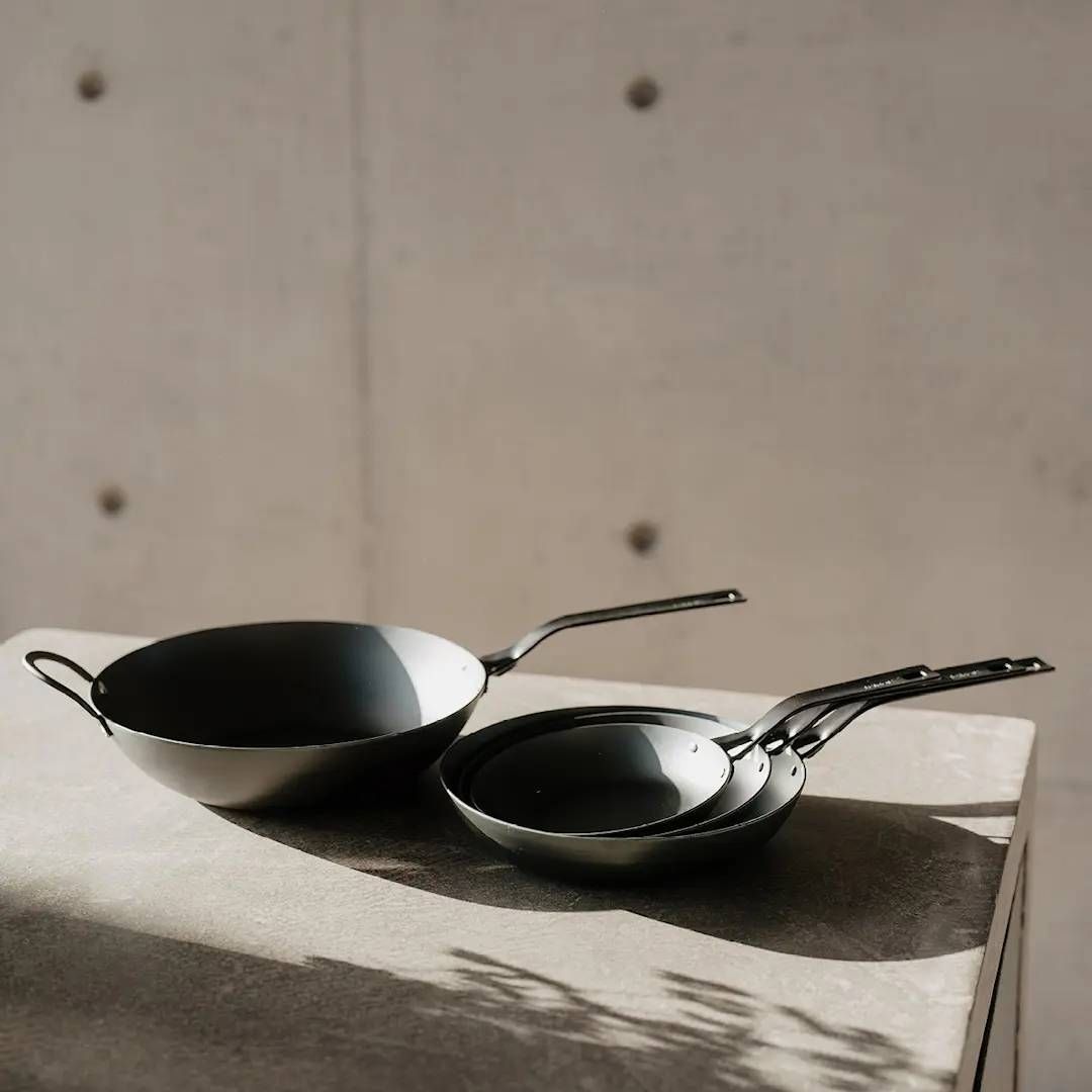 Heirol hiiliteräspannut blacksteel sarjasta pinottu päällekkäin vaalealla pöydällä betoniseinää vasten.