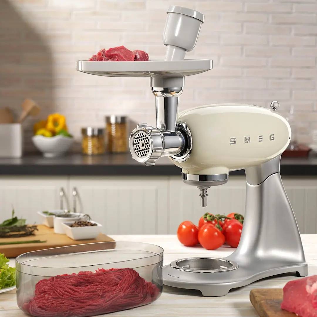 Smeg köttkvarn i krämfärg på köksbänk maler färskt kött med grönsaker i bakgrunden.